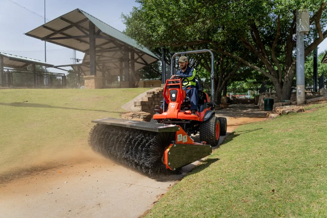 An orange utility vehicle sweeps a dusty path with a front-mounted brush attachment.