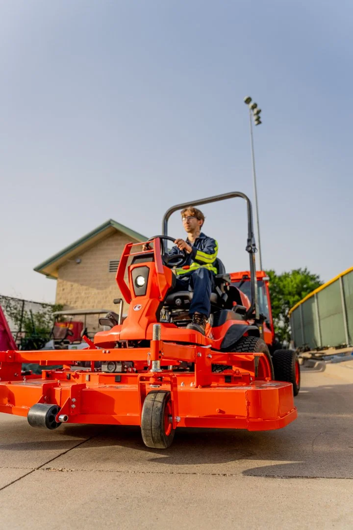 A man sits at the controls of an orange riding mower outside a residential building.