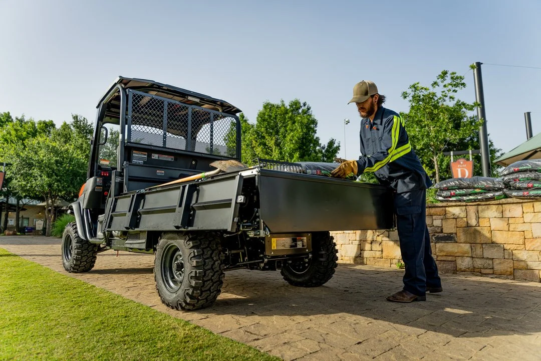 A worker reaches into the cargo bed of a utility vehicle in a sunny outdoor scene.