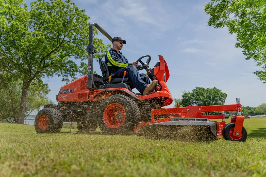 An operator drives an orange tractor towing a mower across a grassy field for equipment video production.