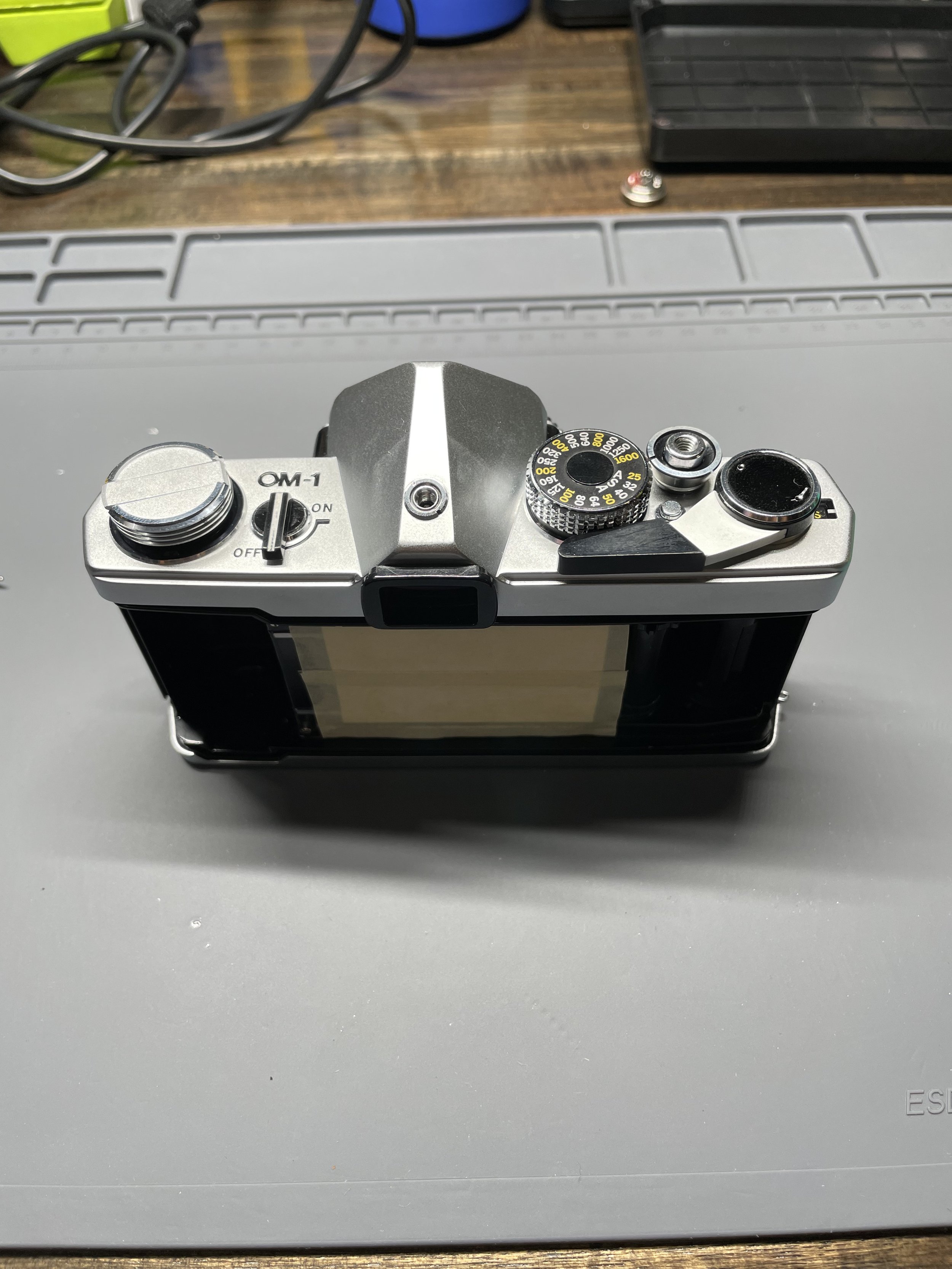An Olympus OM-1 Camera on a workbench, the film back door is removed and masking tape protects the shutter