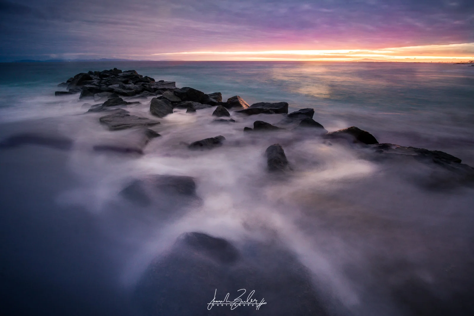 long exposure photograph with a rock on the seashore and misty water flowing over it with a blue sky and teal ocean Captured at Newport Beach pier with the Sony alpha camera