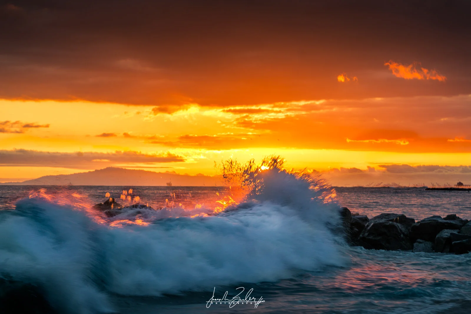vibrant orange sunset with wave crashing on jetty in newport beach california