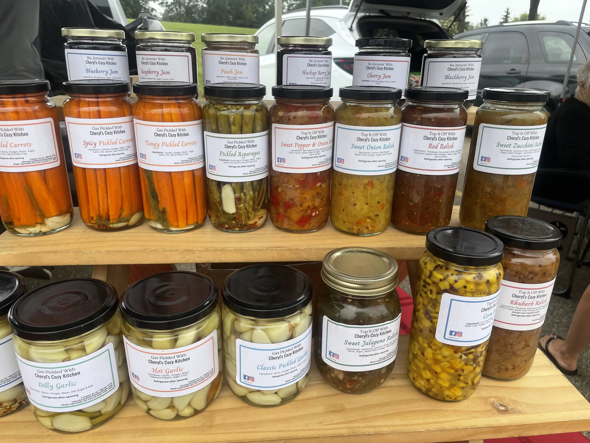 Assortment of homemade pickled vegetables and jams on wooden display shelves at an outdoor market. The jars include carrot pickles, raspberry, peach, blueberry, strawberry, cherry, blackberry jams, and pickled asparagus, peppers, onions, zucchini, jalapeños, corn, garlic, and rhubarb relishes.