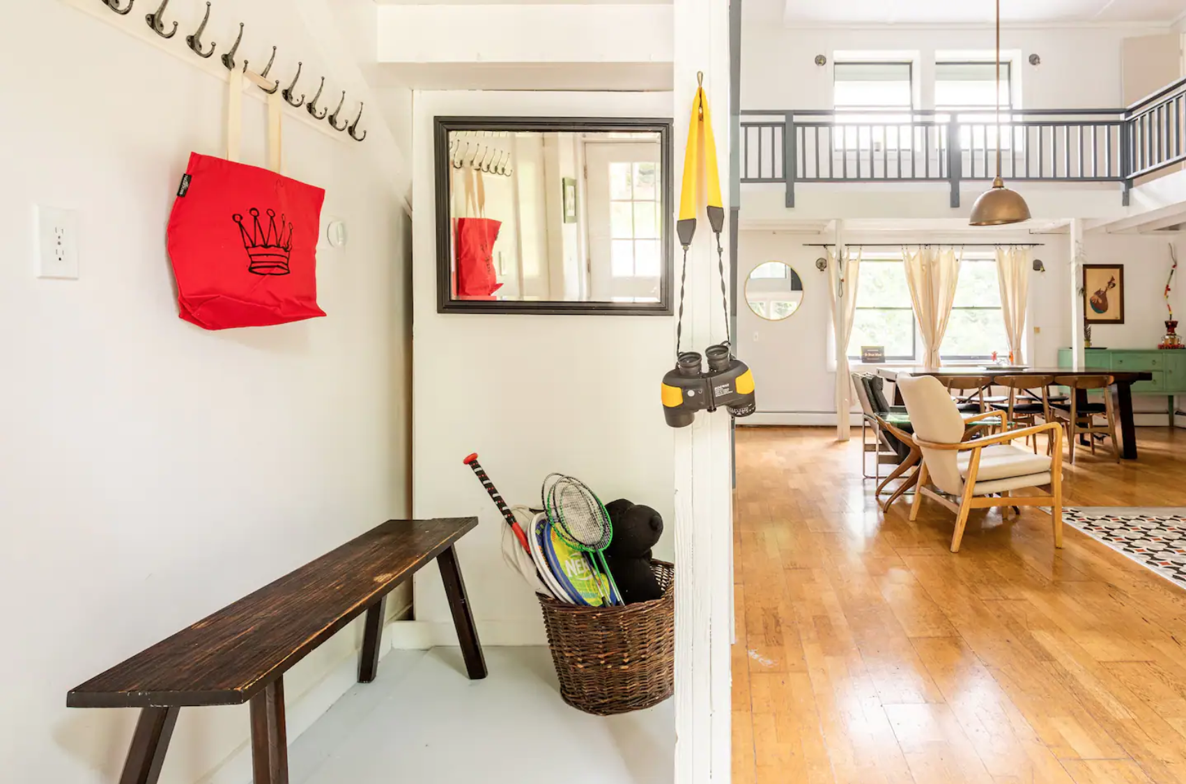 Interior view of a bright room with wooden flooring, featuring a section with a bench, coat hooks, a red bag, and sports equipment in a basket. A mirror and binoculars hang on the wall, while the background reveals a dining area with a table, chairs,
