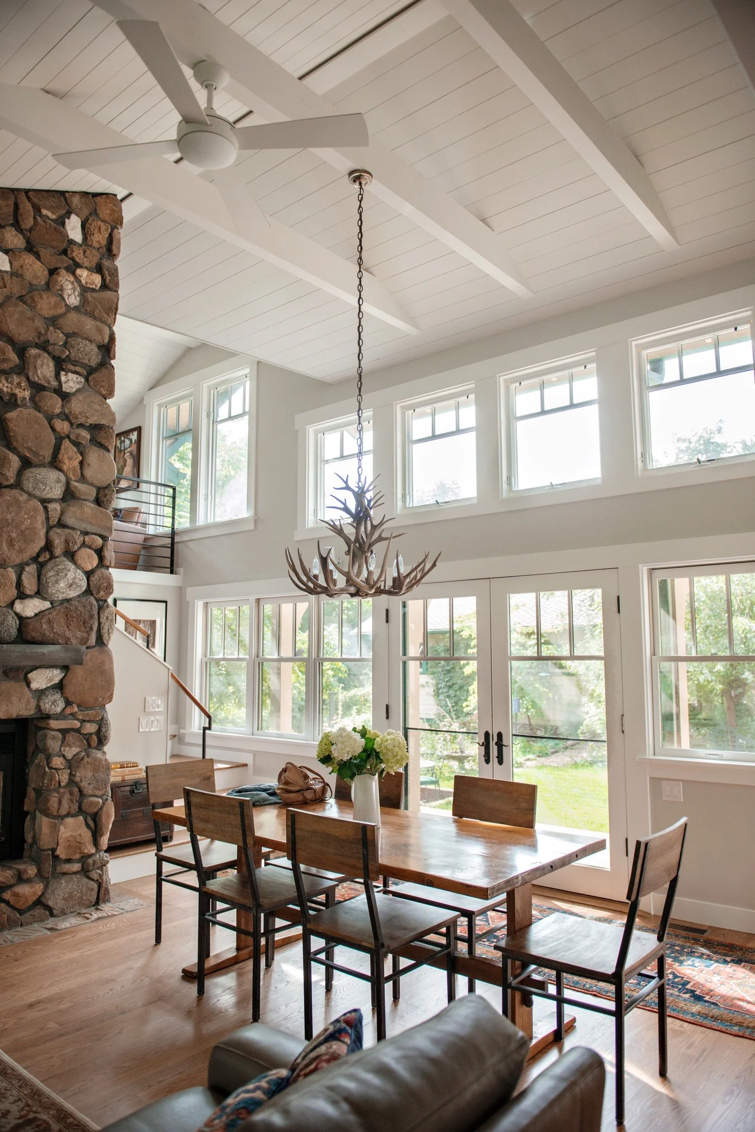 Saltwolf designed dining room with a large wooden table, six chairs, a vase with white flowers, a stone fireplace, vaulted ceiling with white wood panels, and multiple windows allowing natural light to enter.