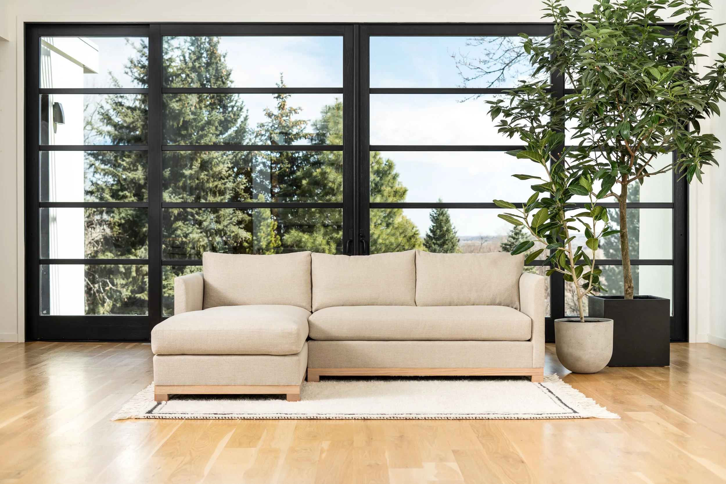 Living room with beige linen Saltwolf Furniture Mapleton sectional sofa, large potted plant, and floor-to-ceiling window with view of trees outside.