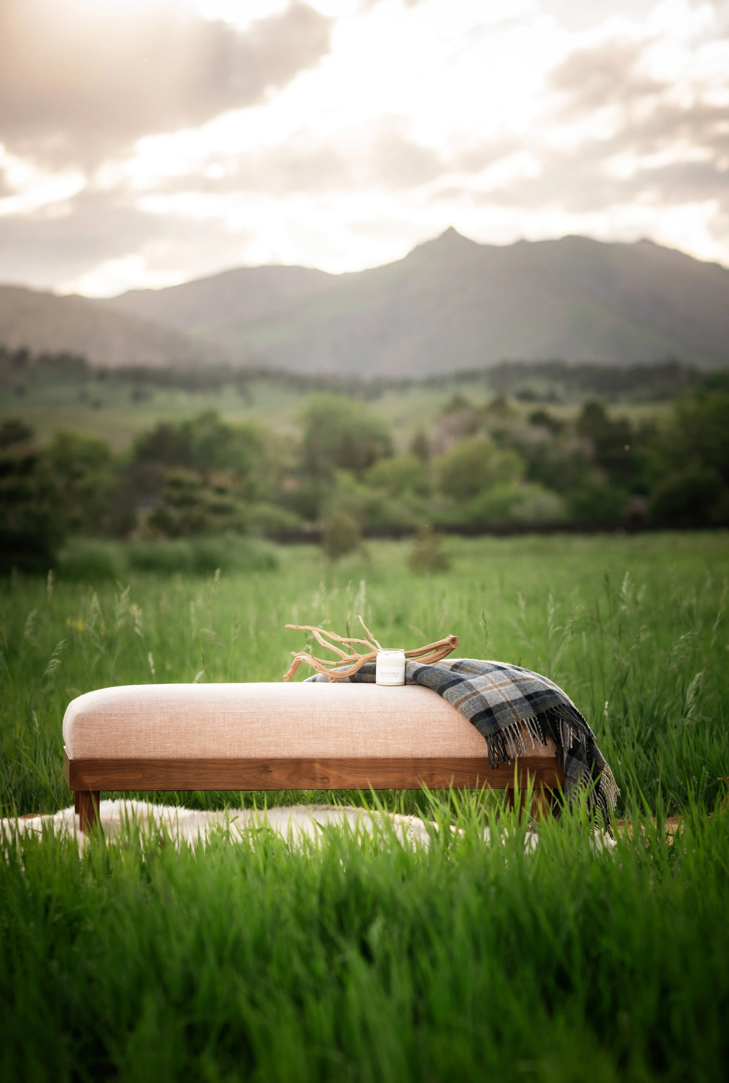 A Saltwolf Furniture Norwood bench in blush linen with maple framer and legs, draped with a plaid blanket, on a grassy field with mountains and cloudy sky in the background.