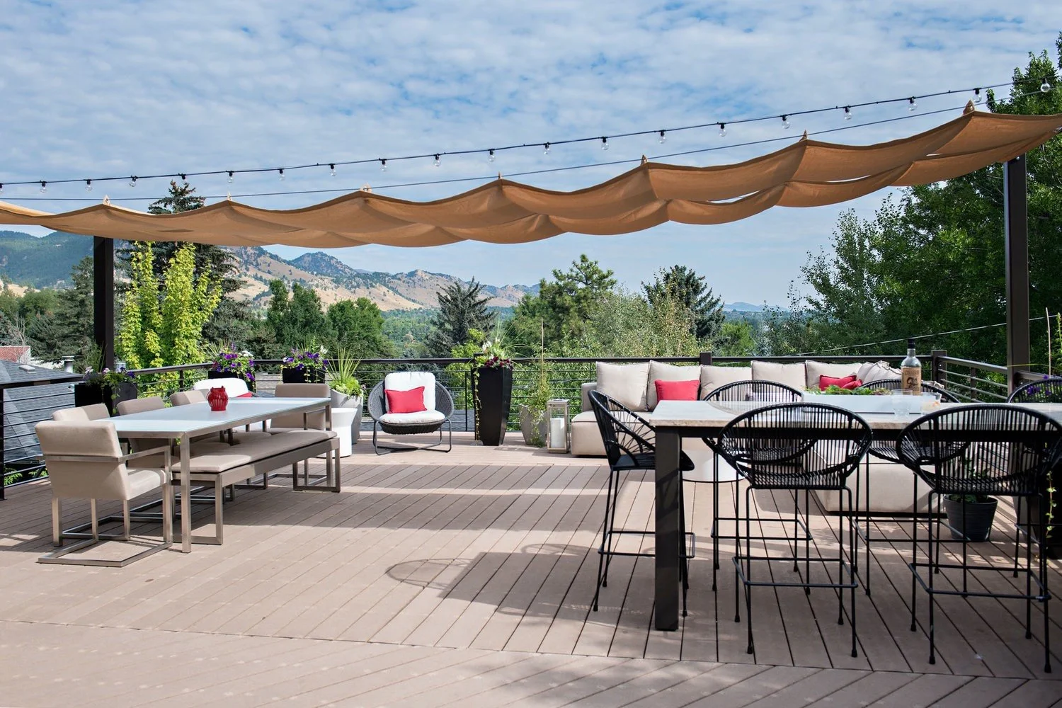 Saltwolf designed outdoor patio with modern furniture, including a white dining table with beige chairs, a black wire bar-height table with black wire chairs, and a beige outdoor sofa with red cushions.