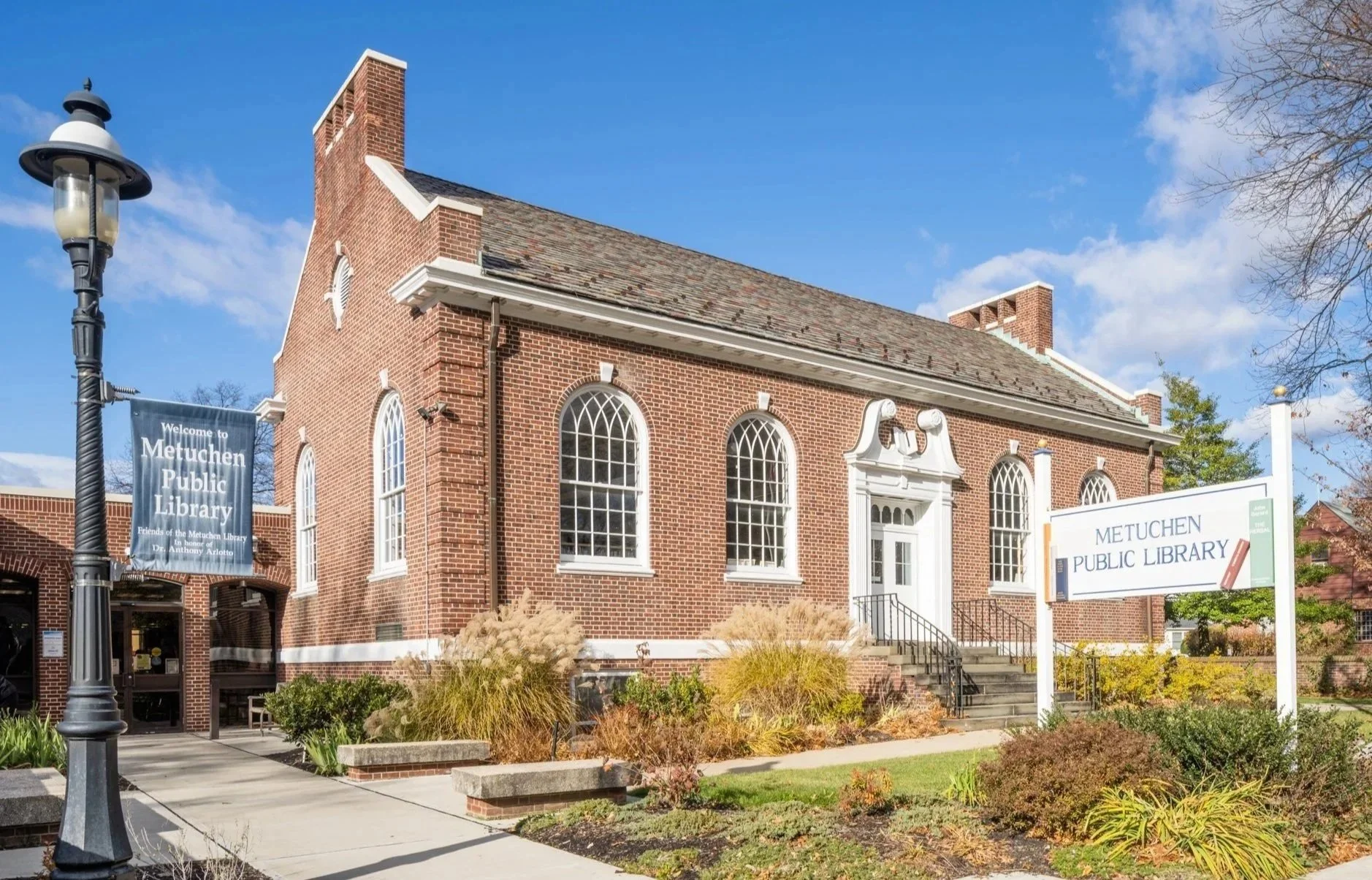 Exterior view of Metuchen Public Library building with brick facade, arched windows, a sign indicating the library's name, and landscaping with bushes and plants under a blue sky.