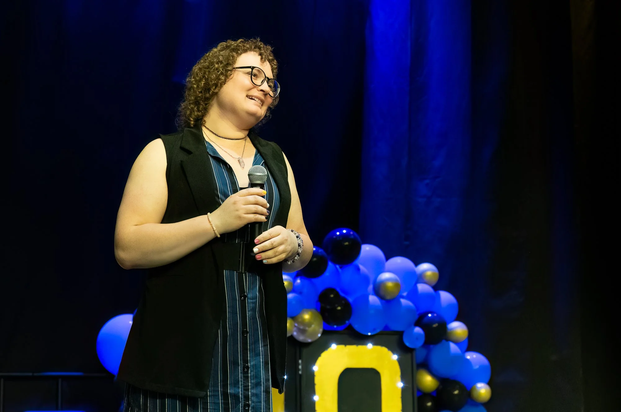 A smiling woman with curly hair wearing a blue dress with a suit vest holding a microphone on a stage with blue and gold accents.