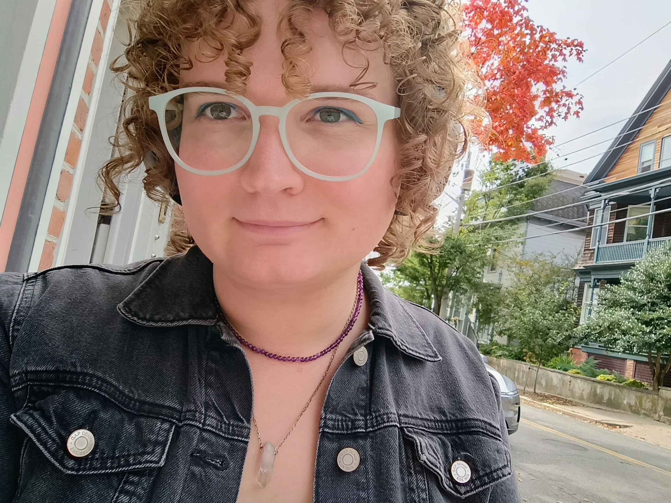A woman with curly blonde hair and glasses taking a selfie outdoors on a cloudy day, with autumn trees and residential buildings in the background.