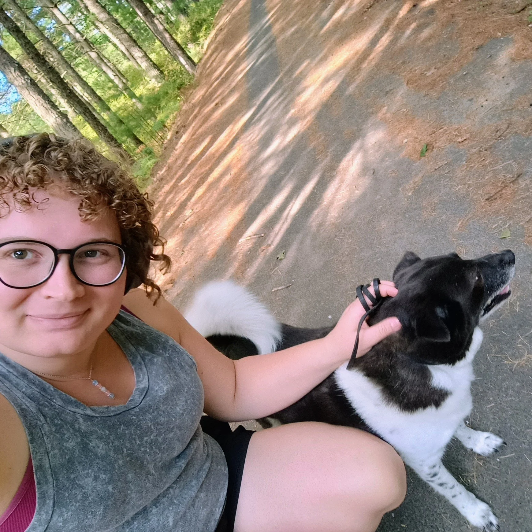 A person with curly hair and glasses smiling for a selfie outdoors in a wooded area, sitting on the ground with a black and white dog, holding the dog's collar, both enjoying a sunny day.