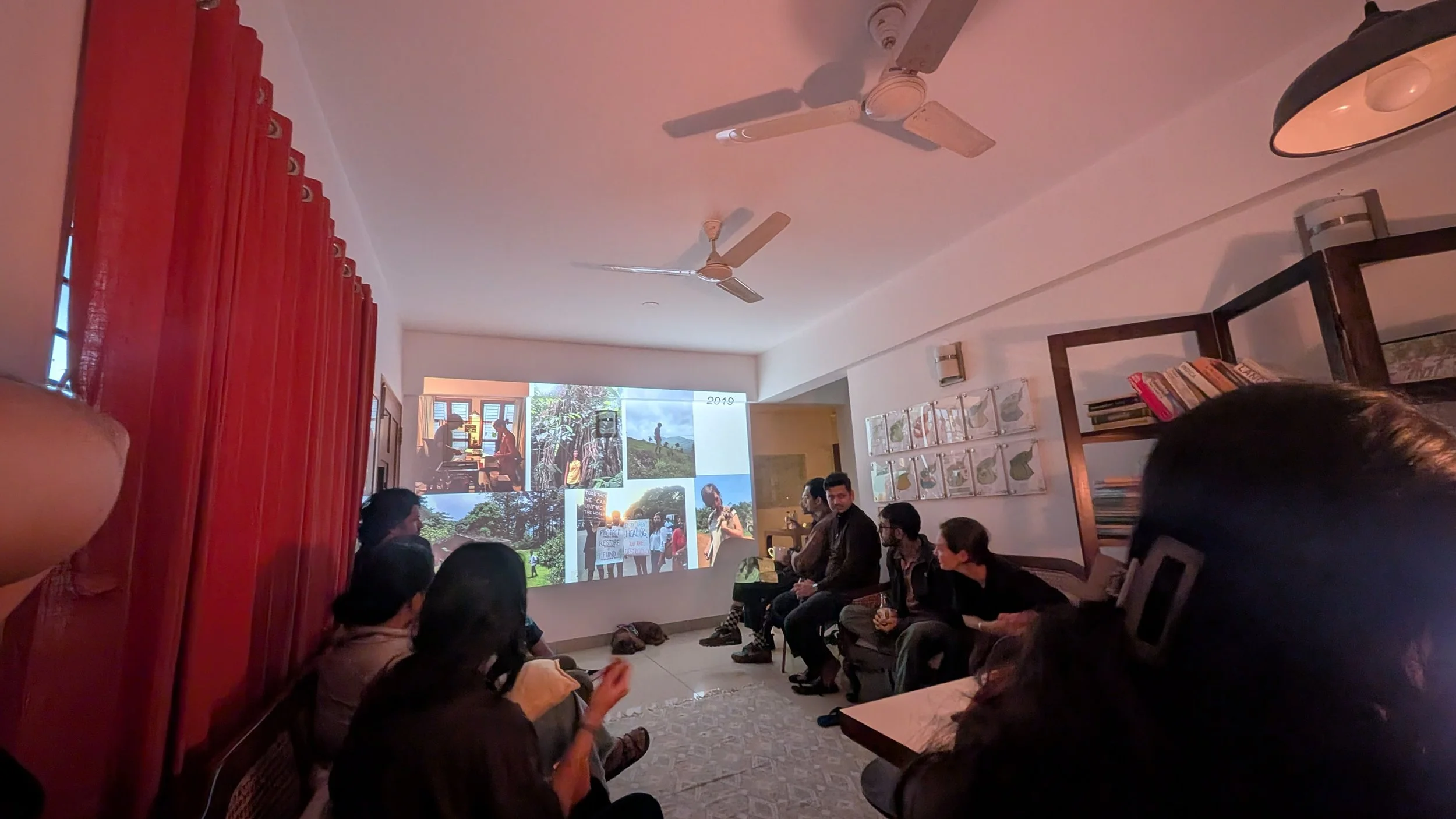 People sitting in a room watching a presentation projected on the wall, featuring a collage of outdoor photos with trees, mountains, and people hiking or exploring nature.