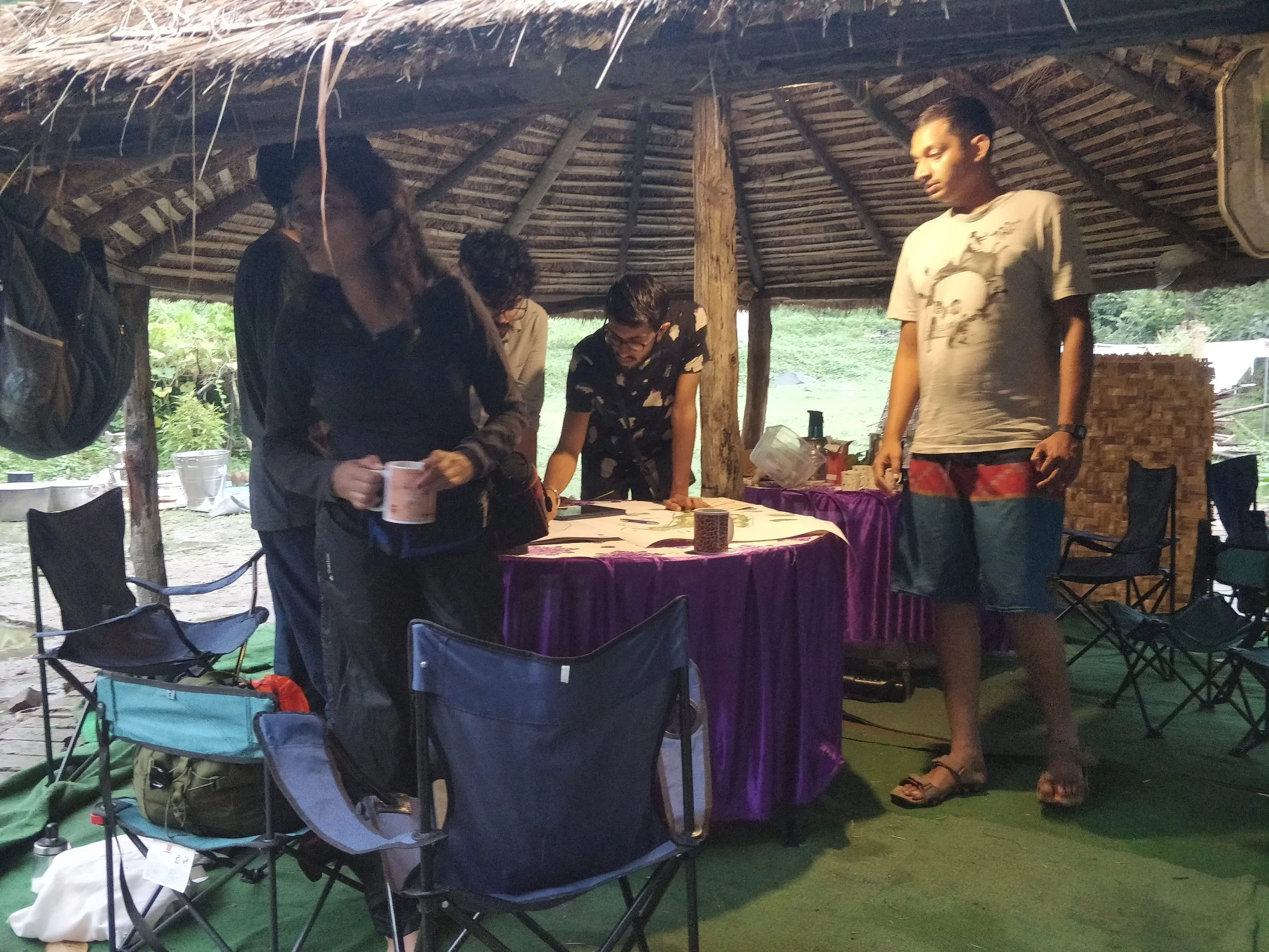 Group of five people standing around a round table with purple tablecloth under a thatched roof structure, with outdoor greenery visible in the background. One person is holding a cup, others are looking at items on the table.