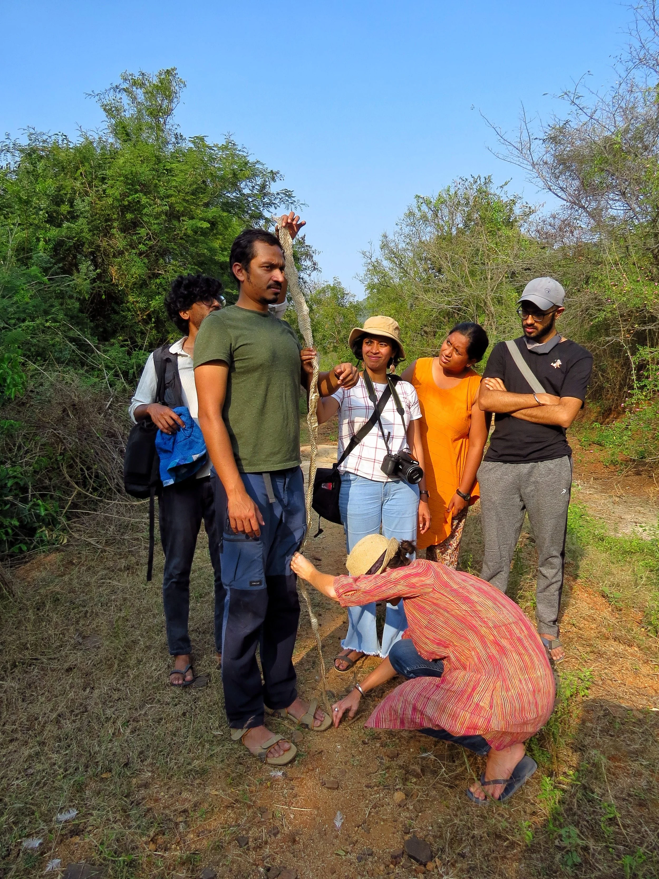 A group of people standing on a dirt path in nature, with one person squatting and touching the ground, surrounded by bushes and trees, under a clear blue sky.