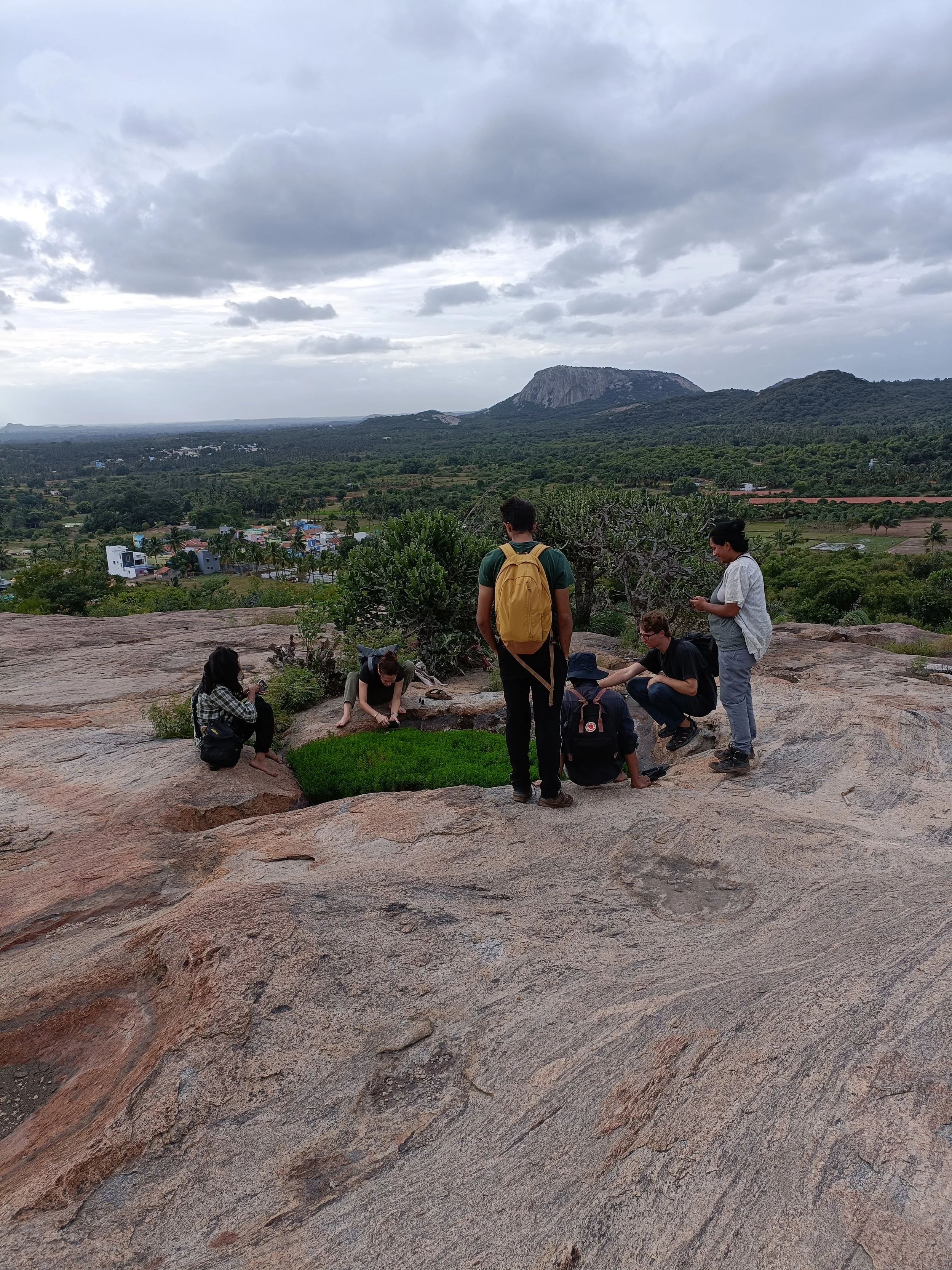Group of people exploring a rocky hilltop with lush greenery and distant mountains under a cloudy sky.