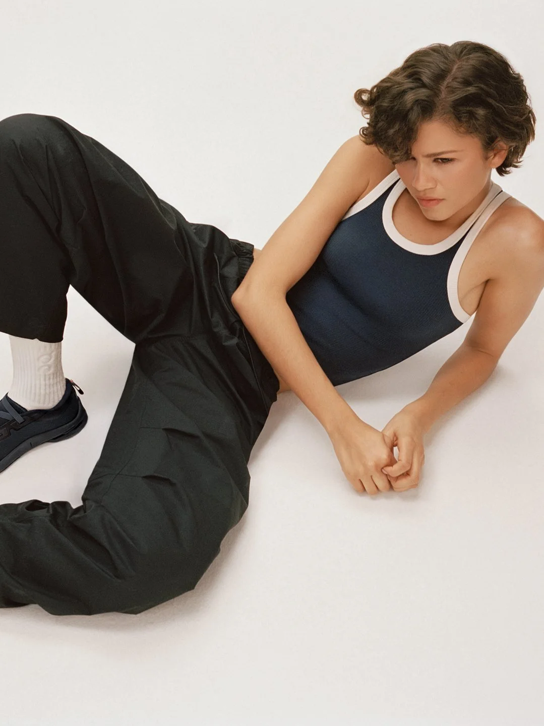  Zendaya reclining against a white studio backdrop in a navy tank top, black trousers and On sneakers, photographed by Sean Thomas in London for On. 