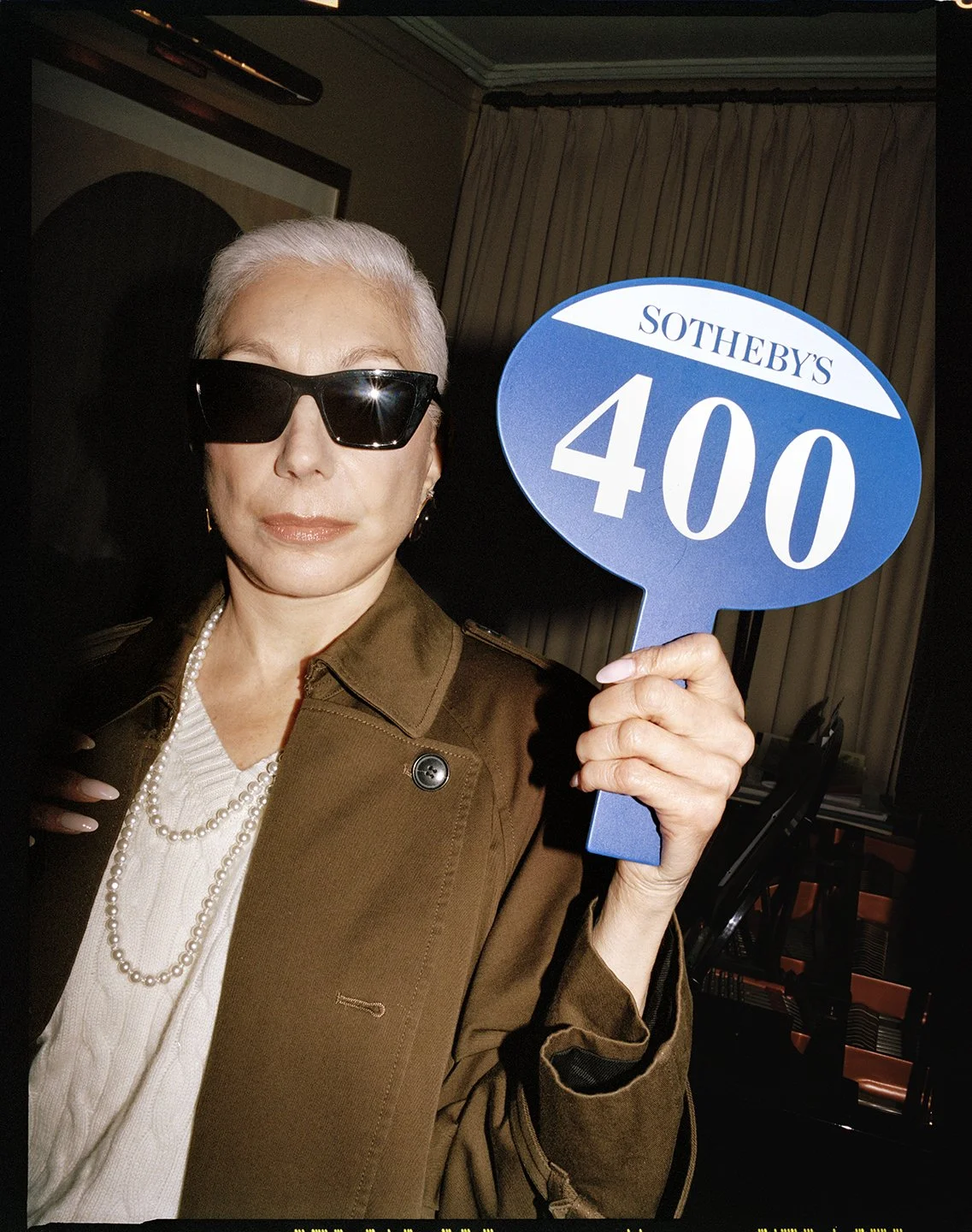  Portrait of a woman in black sunglasses and a brown trench coat holding a blue Sotheby’s auction paddle, photographed by Sean Thomas. 