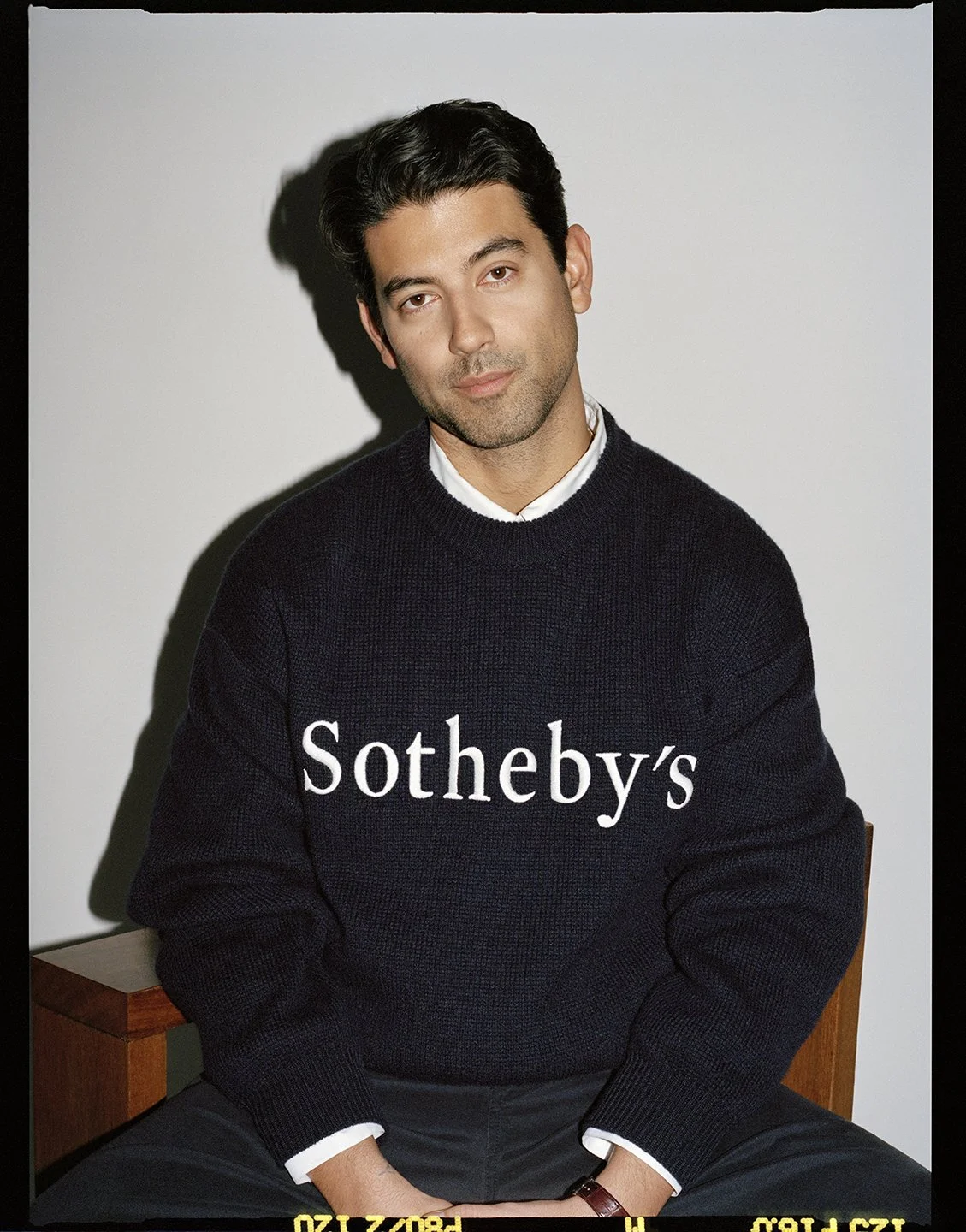  Portrait of Sotheby’s specialist Ashkan Baghestani seated in a navy Sotheby’s sweater, photographed by Sean Thomas for the FRAME collaboration. 