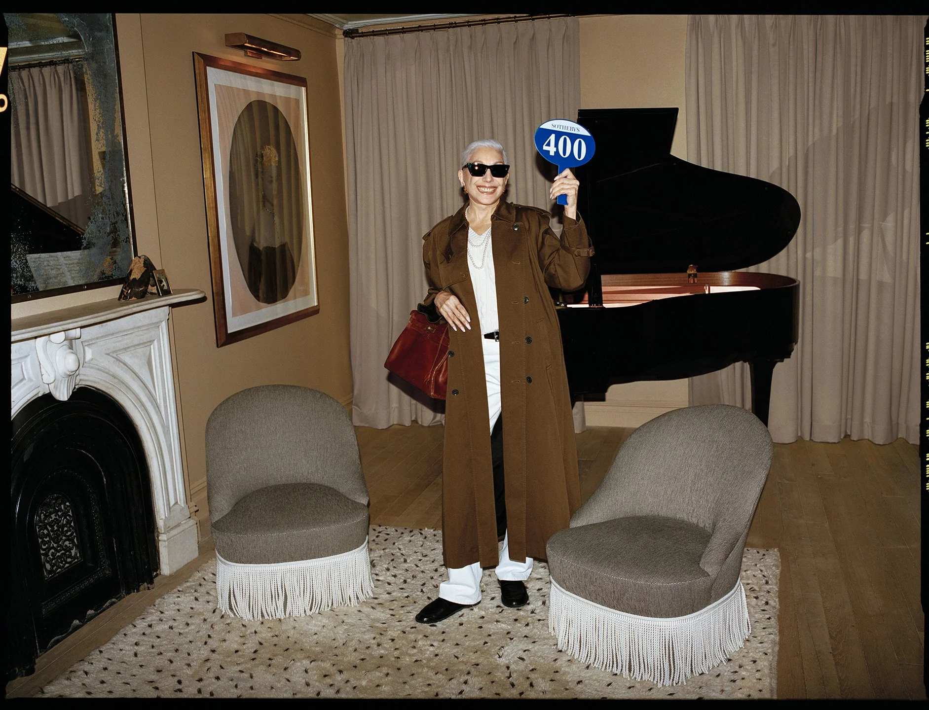  Full-length portrait of a woman in a brown trench coat and sunglasses holding a Sotheby’s auction paddle beside a grand piano. 
