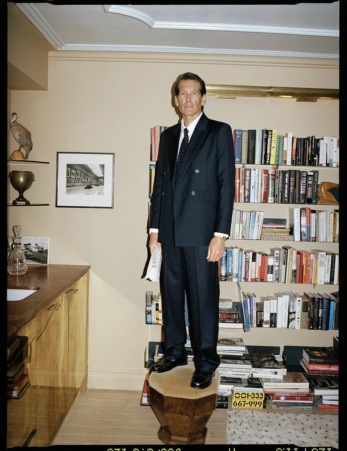  Portrait of a man in a double-breasted navy suit standing before bookshelves, photographed by Sean Thomas for FRAME x Sotheby’s. 