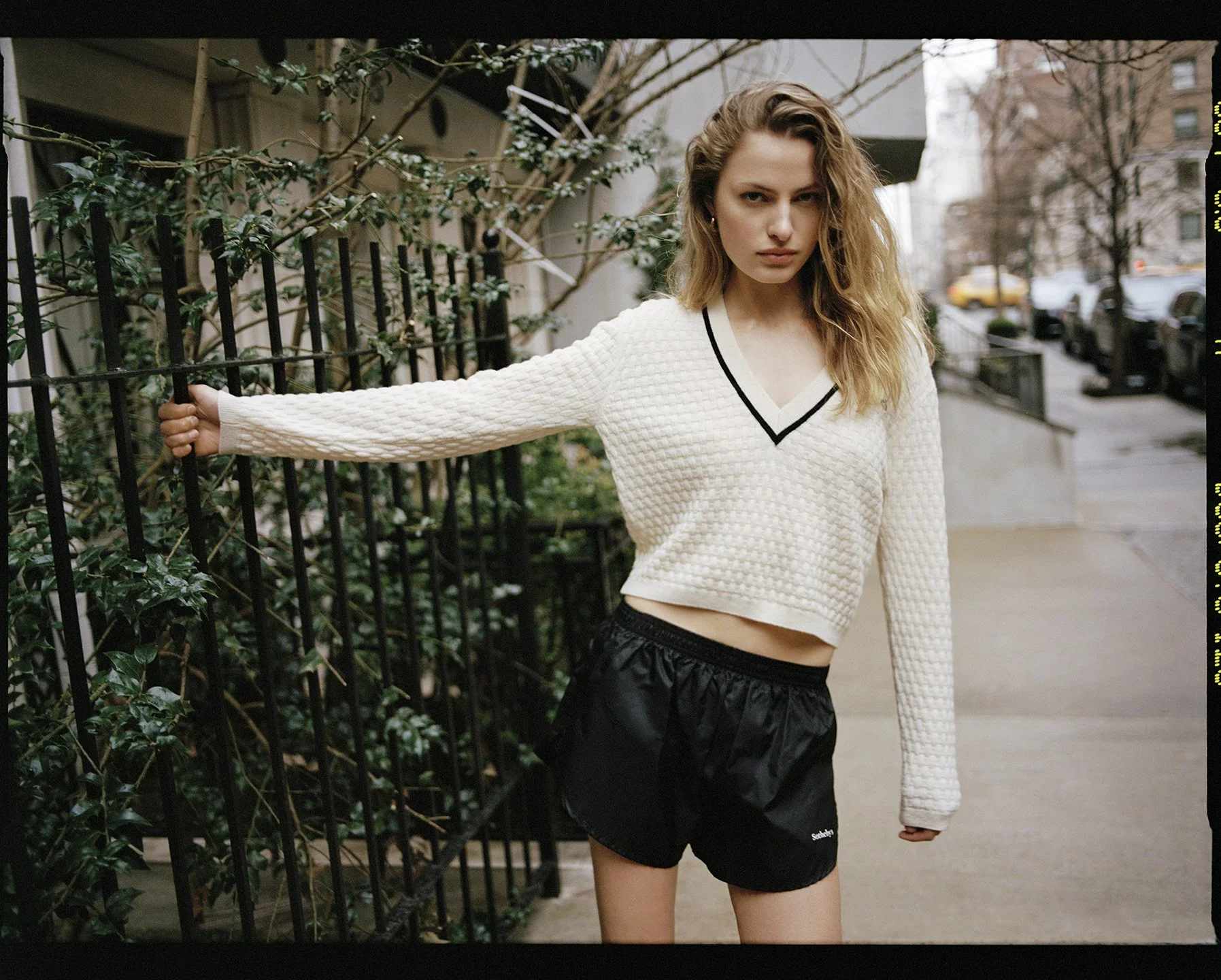  Felice Nova Noordhoff outdoors in New York wearing a white V-neck sweater and black Sotheby’s shorts, photographed by Sean Thomas. 