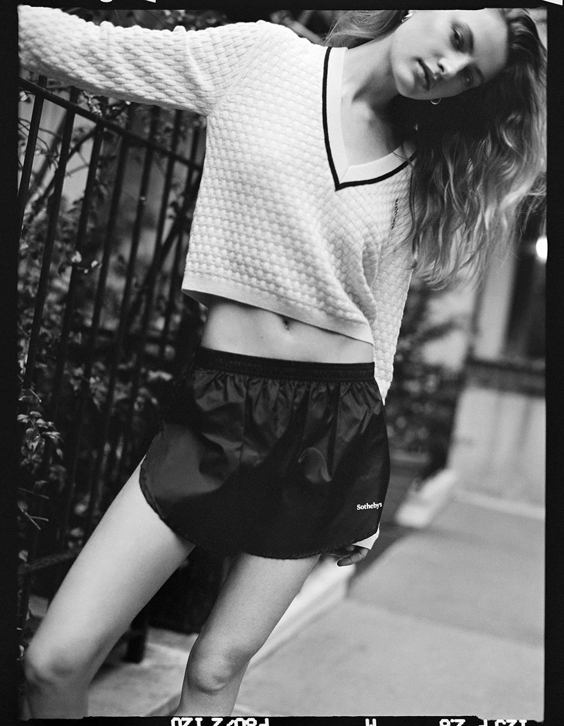  Black-and-white portrait of Felice Nova Noordhoff leaning beside an iron railing in FRAME x Sotheby’s knitwear and shorts. 
