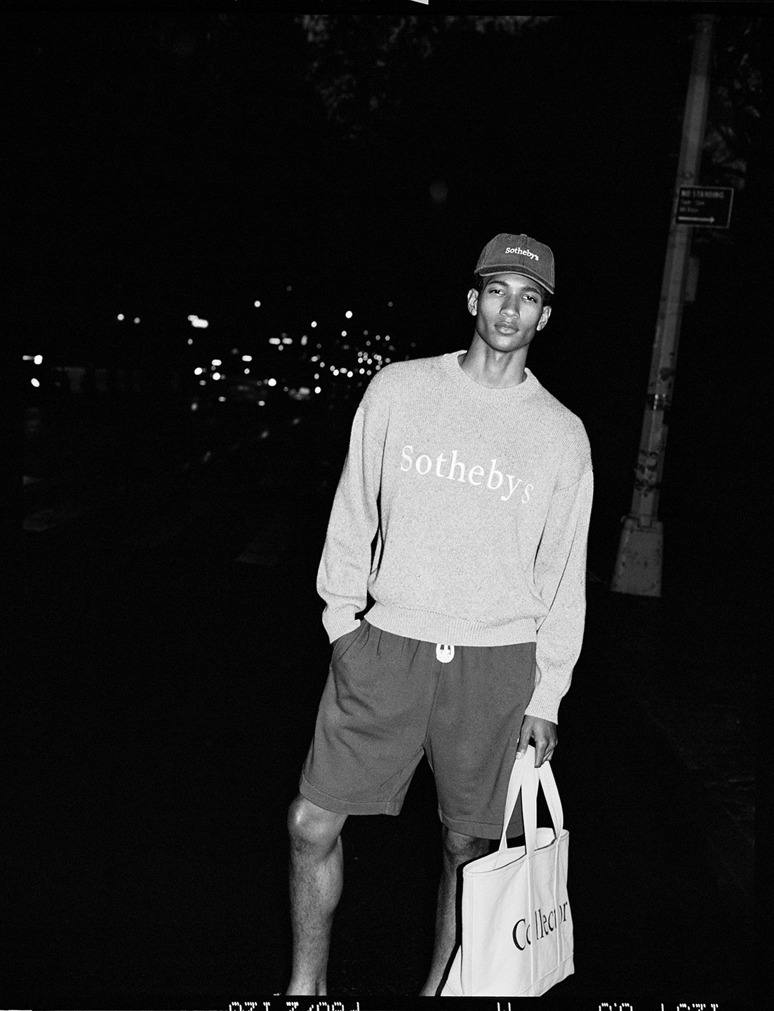  Joshua Seth Williams at night in a Sotheby’s cap and sweatshirt with a Collector tote, photographed in black and white by Sean Thomas. 