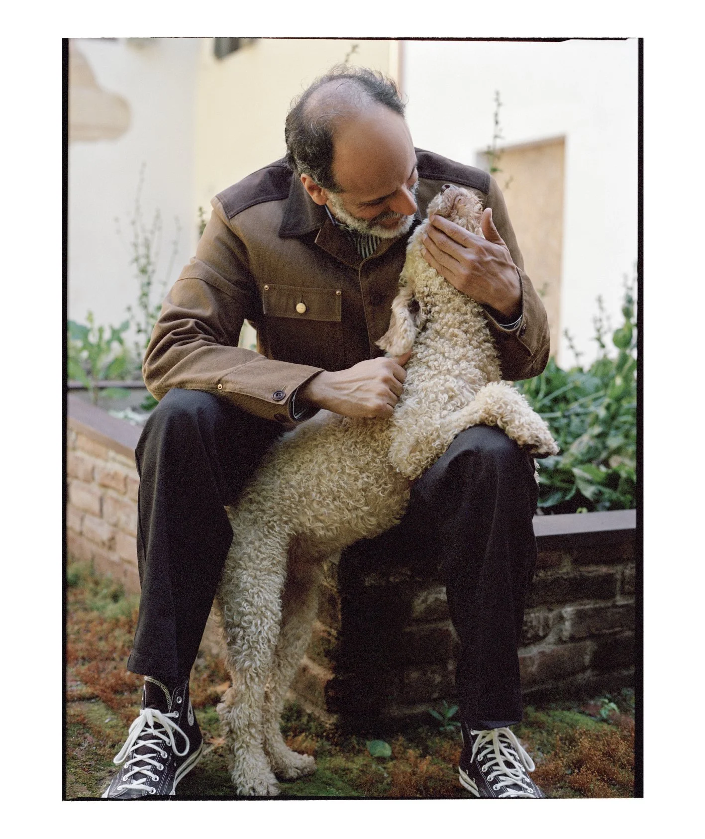  Luca Guadagnino seated outdoors embracing his curly-haired dog in a color portrait photographed by Sean Thomas for Door magazine. 