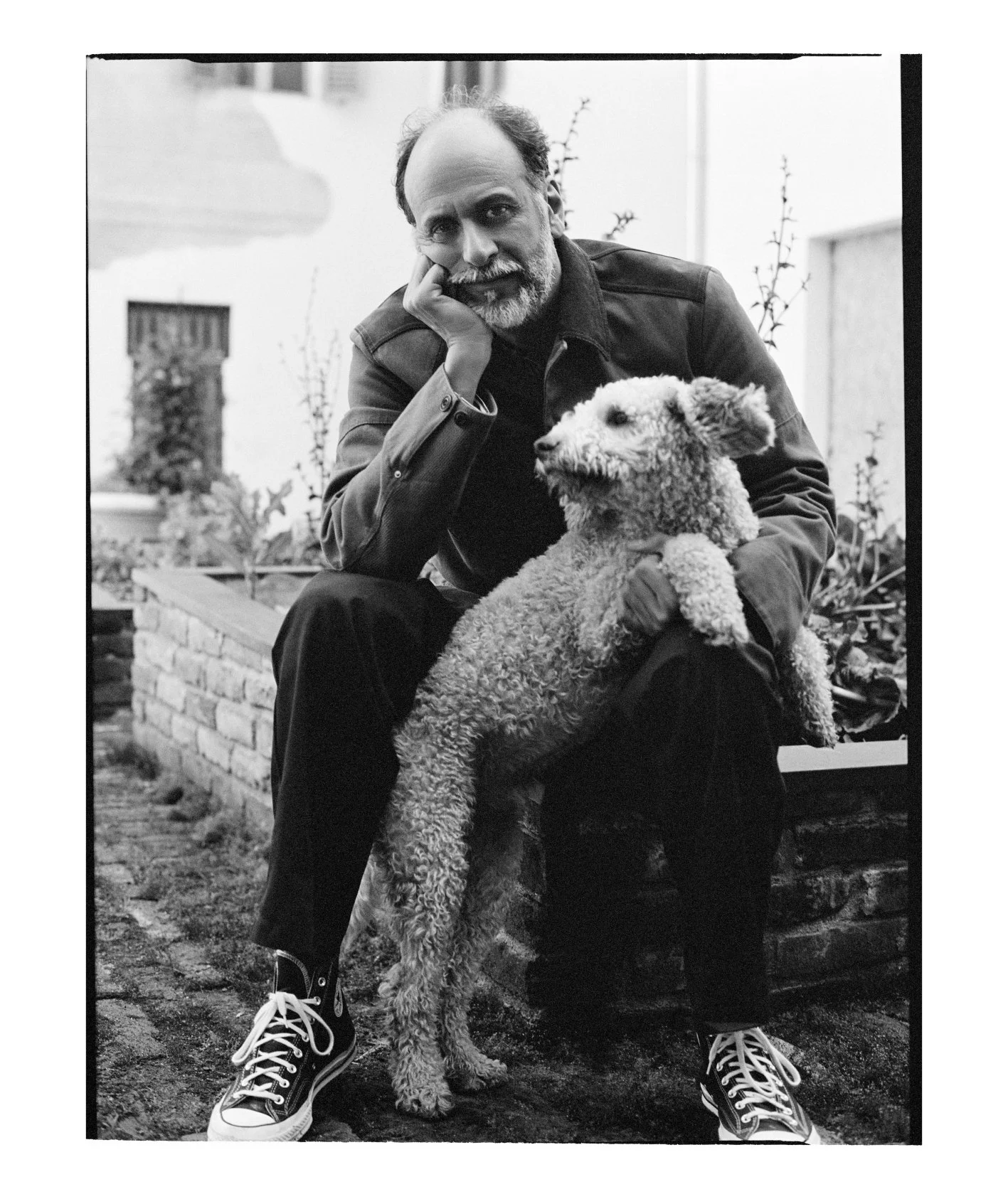  Black-and-white portrait of Luca Guadagnino seated outdoors with his curly-haired dog resting against him, photographed by Sean Thomas for Door magazine. 