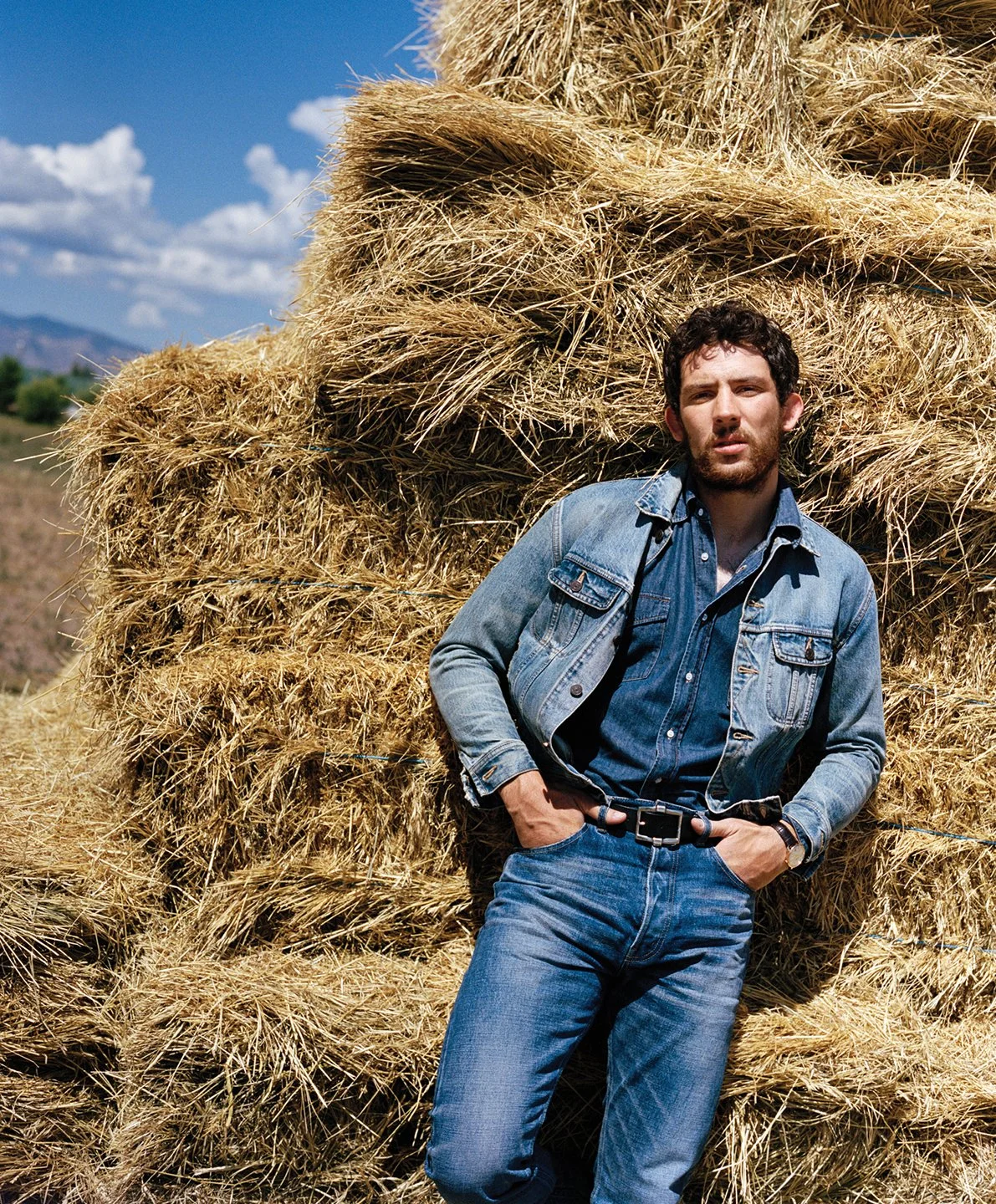  Josh O’Connor partially concealed by stacked hay bales beneath a blue Colorado sky, photographed by Sean Thomas for WSJ. Magazine. 