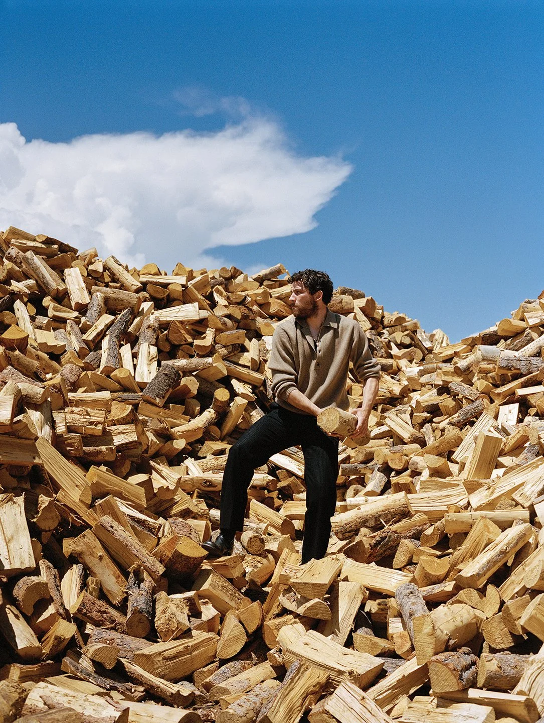  Josh O’Connor holding a log amid a vast woodpile under a cloud-filled sky near Monte Vista, Colorado, photographed by Sean Thomas for WSJ. Magazine. 