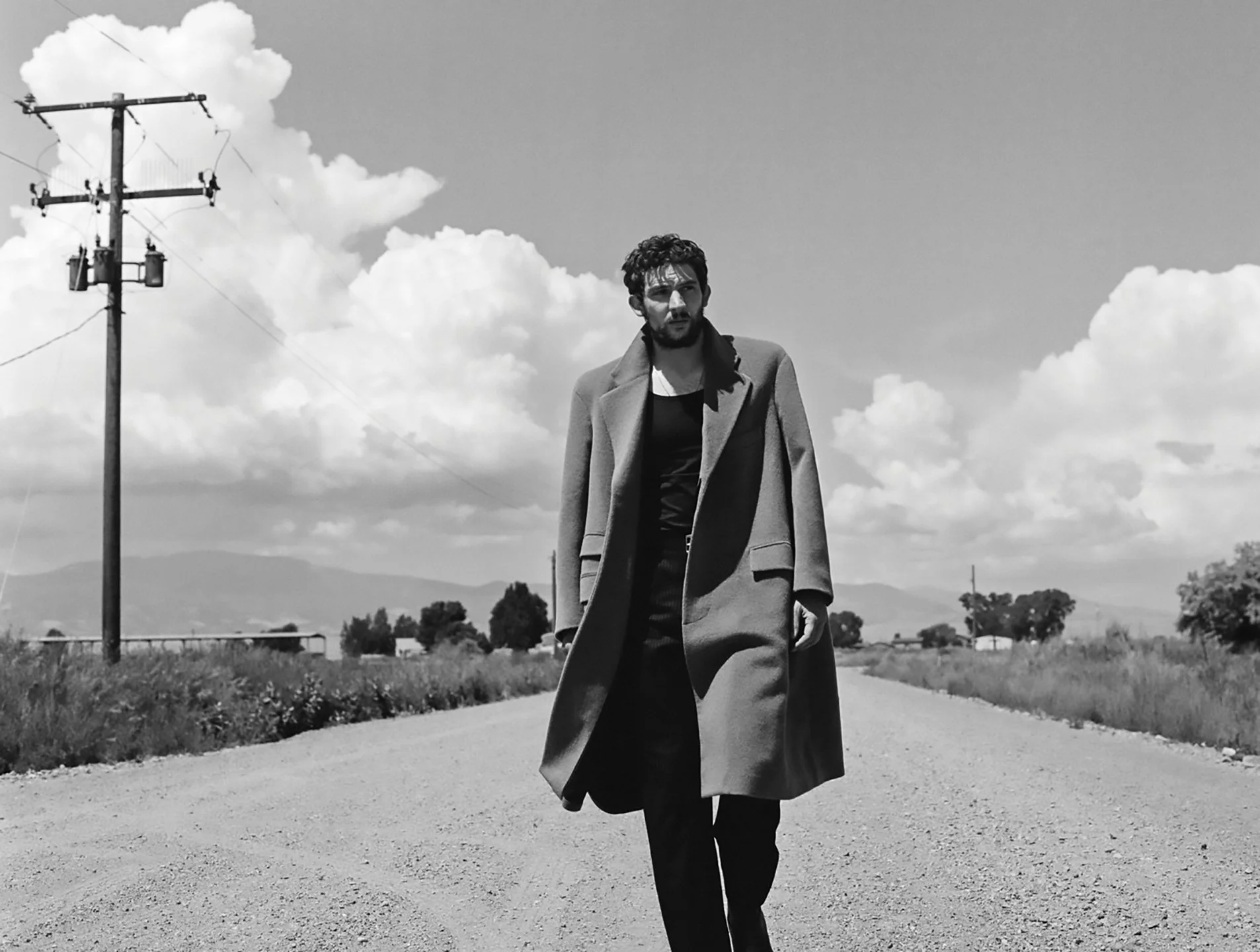  Black-and-white portrait of Josh O’Connor walking along a rural Colorado road in a long overcoat beneath dramatic clouds, photographed by Sean Thomas for WSJ. Magazine. 