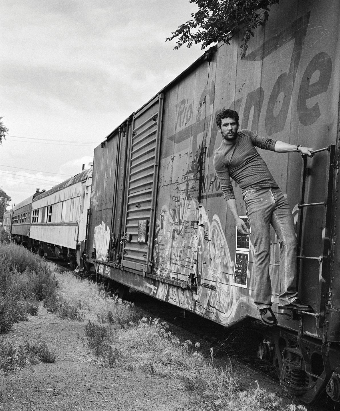  Black-and-white portrait of Josh O’Connor standing on the ladder of a weathered freight train in Colorado, photographed by Sean Thomas for WSJ. Magazine. 