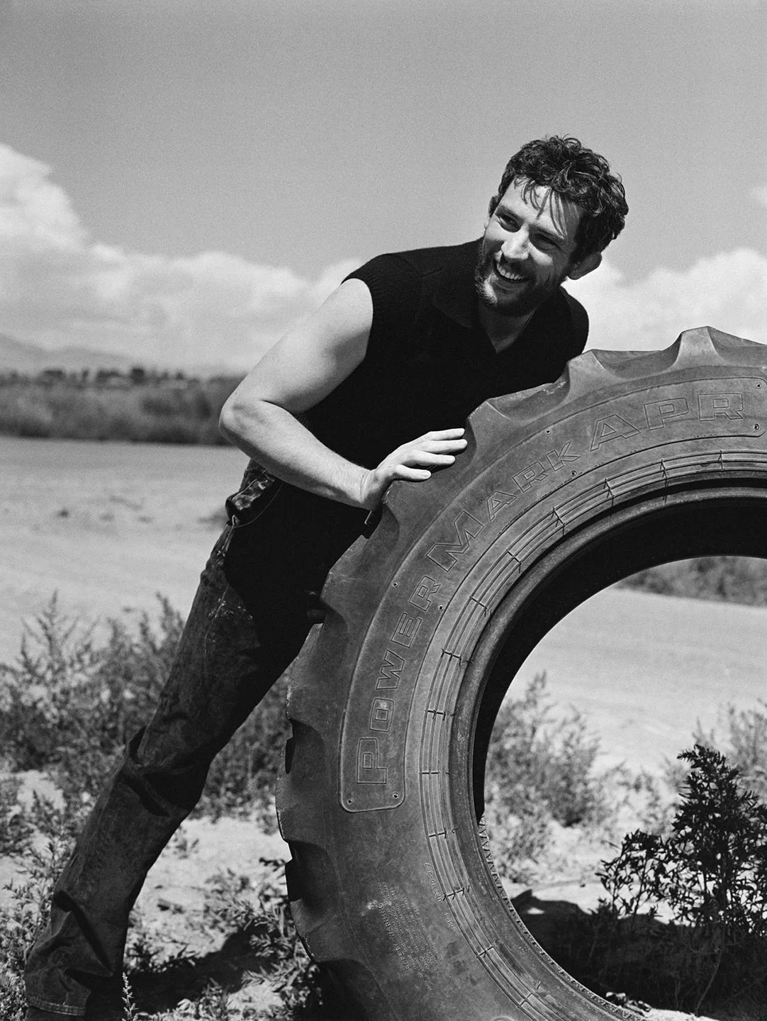  Josh O’Connor laughing beside an enormous tractor tire in rural Colorado, photographed in black and white by Sean Thomas for WSJ. Magazine. 