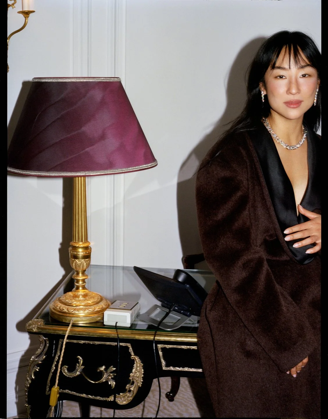 Greta Lee in a dark tailored look with jewelry beside a vintage lamp, photographed by Sean Thomas for Sotheby’s Magazine and styled by Geraldine Saglio. 
