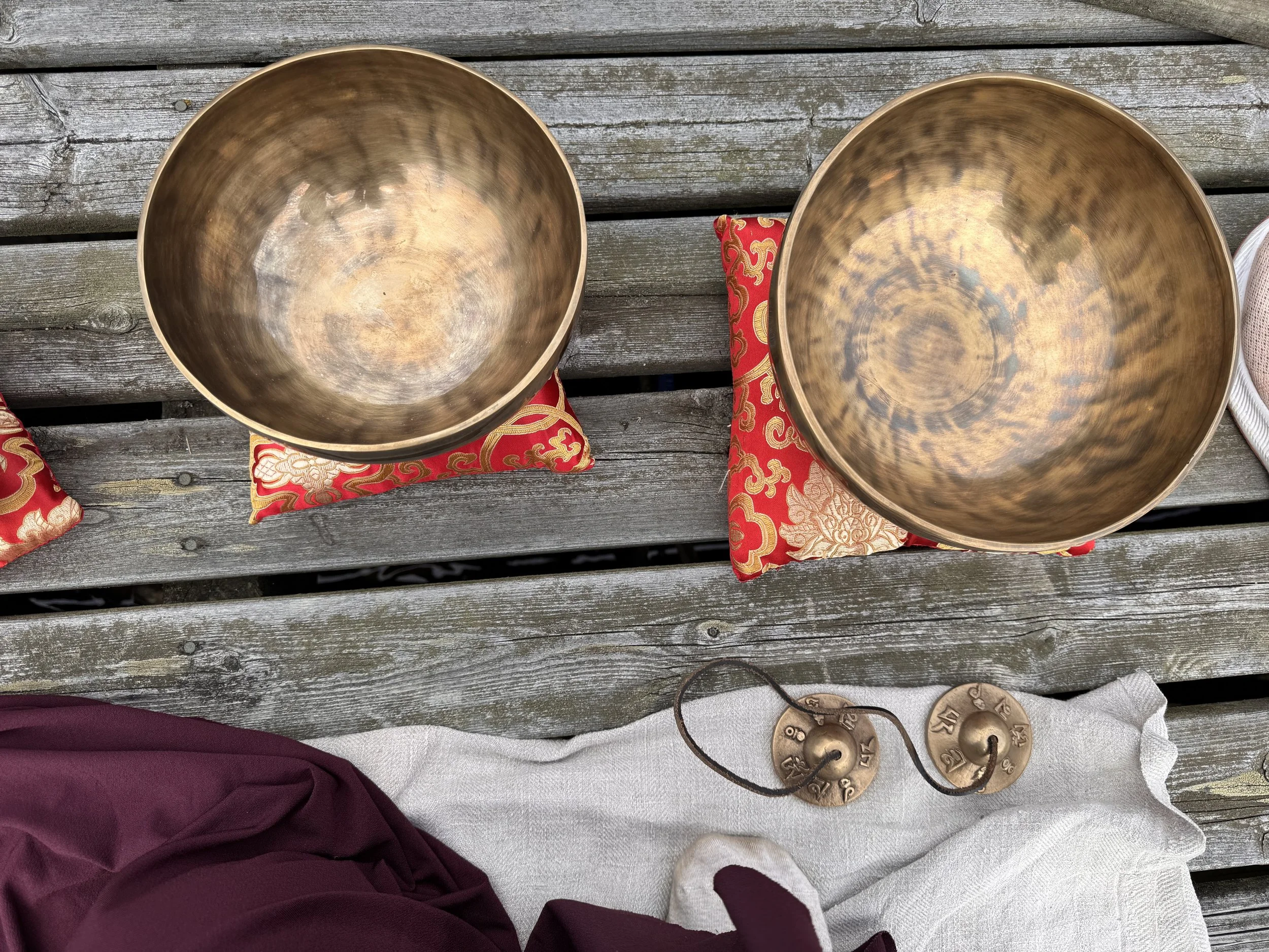 Two singing bowls resting on decorative red and gold cushions on a wooden outdoor surface, with a white cloth underneath and coins nearby.