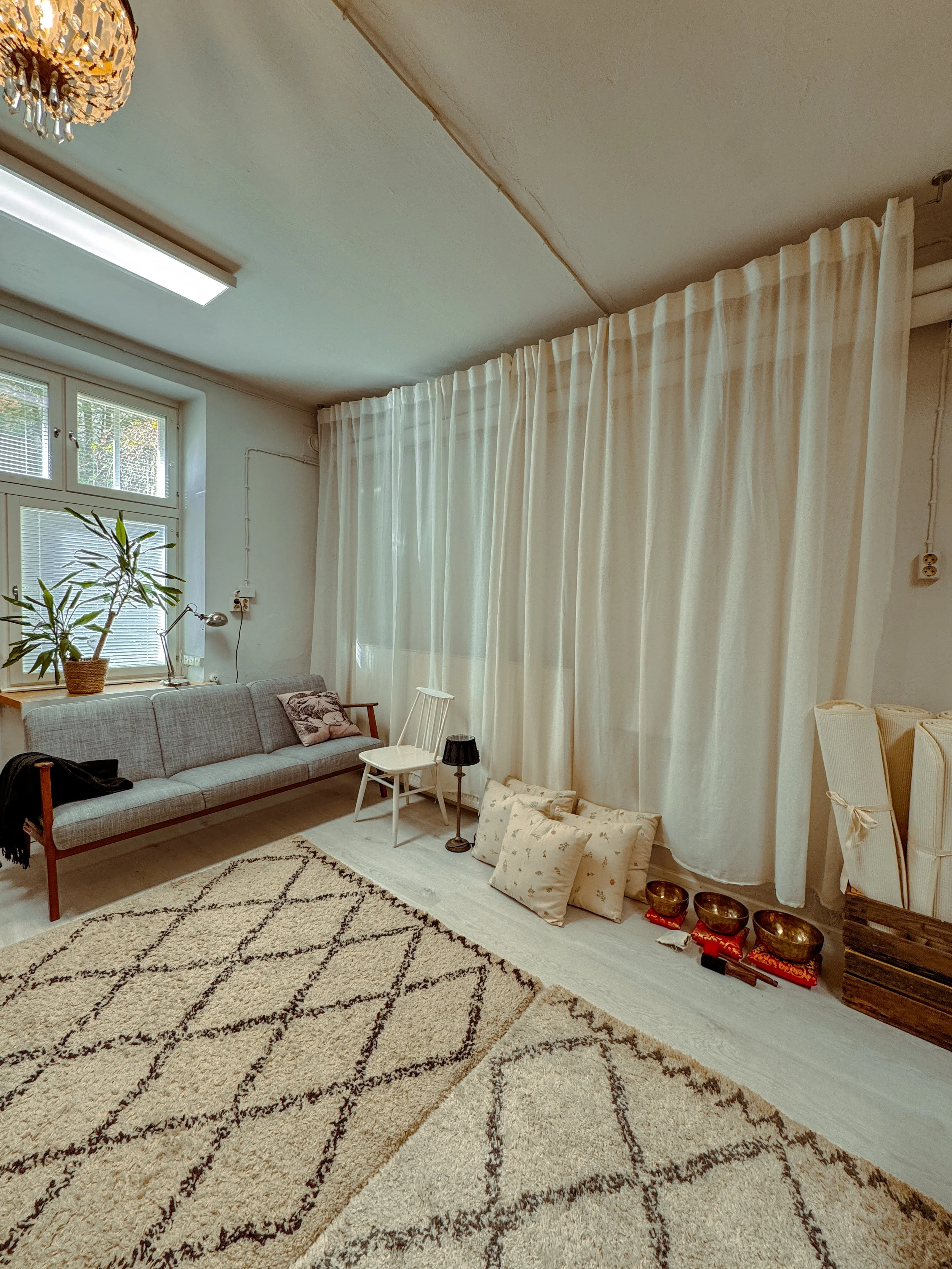 A cozy living room with beige and white curtains, a gray sofa with pillows, a potted plant by the window, a small white chair, a black lampshade, a rug with geometric patterns, and a row of golden bowls and red drums on the floor near the wall.