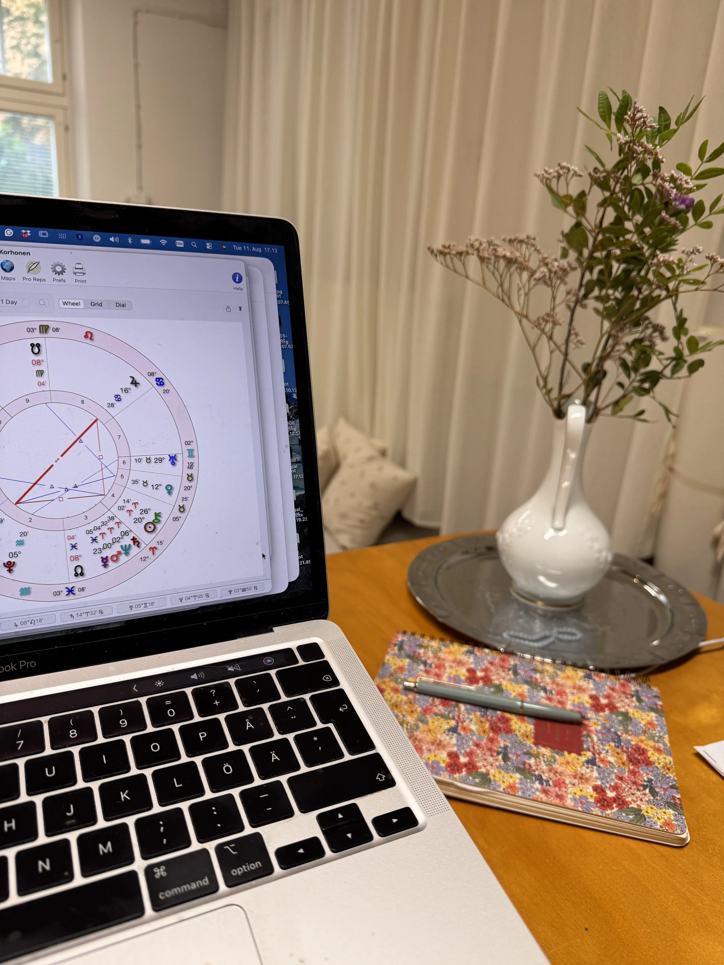 Close-up view of a laptop keyboard, a colorful floral notebook, a silver pen, a glass vase with flowers, and a silver tray on a wooden table.
