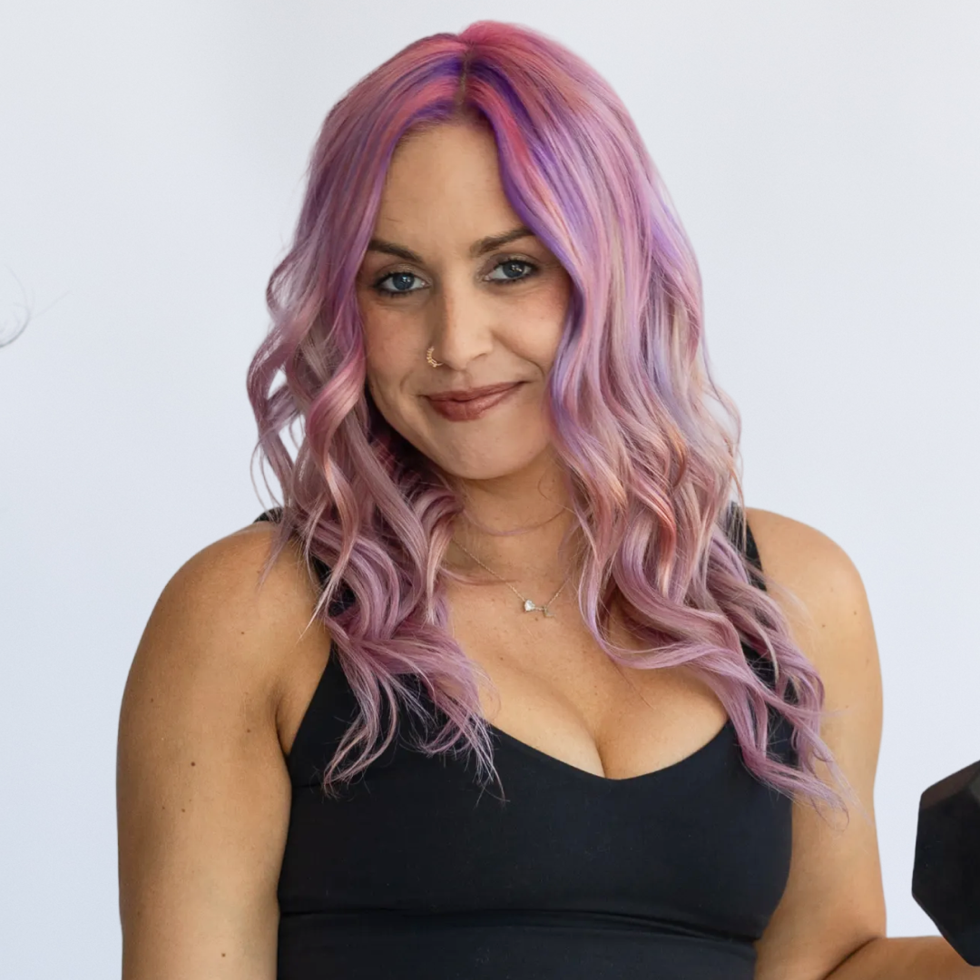A woman with pink and purple wavy hair holding a dumbbell, standing against a plain white background.
