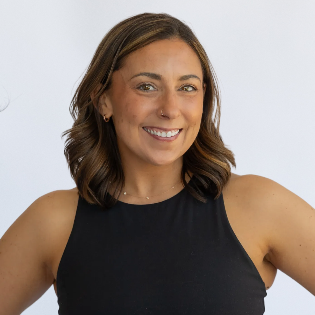 Smiling woman with long wavy hair, wearing a black sleeveless top and jewelry, standing against a plain background.