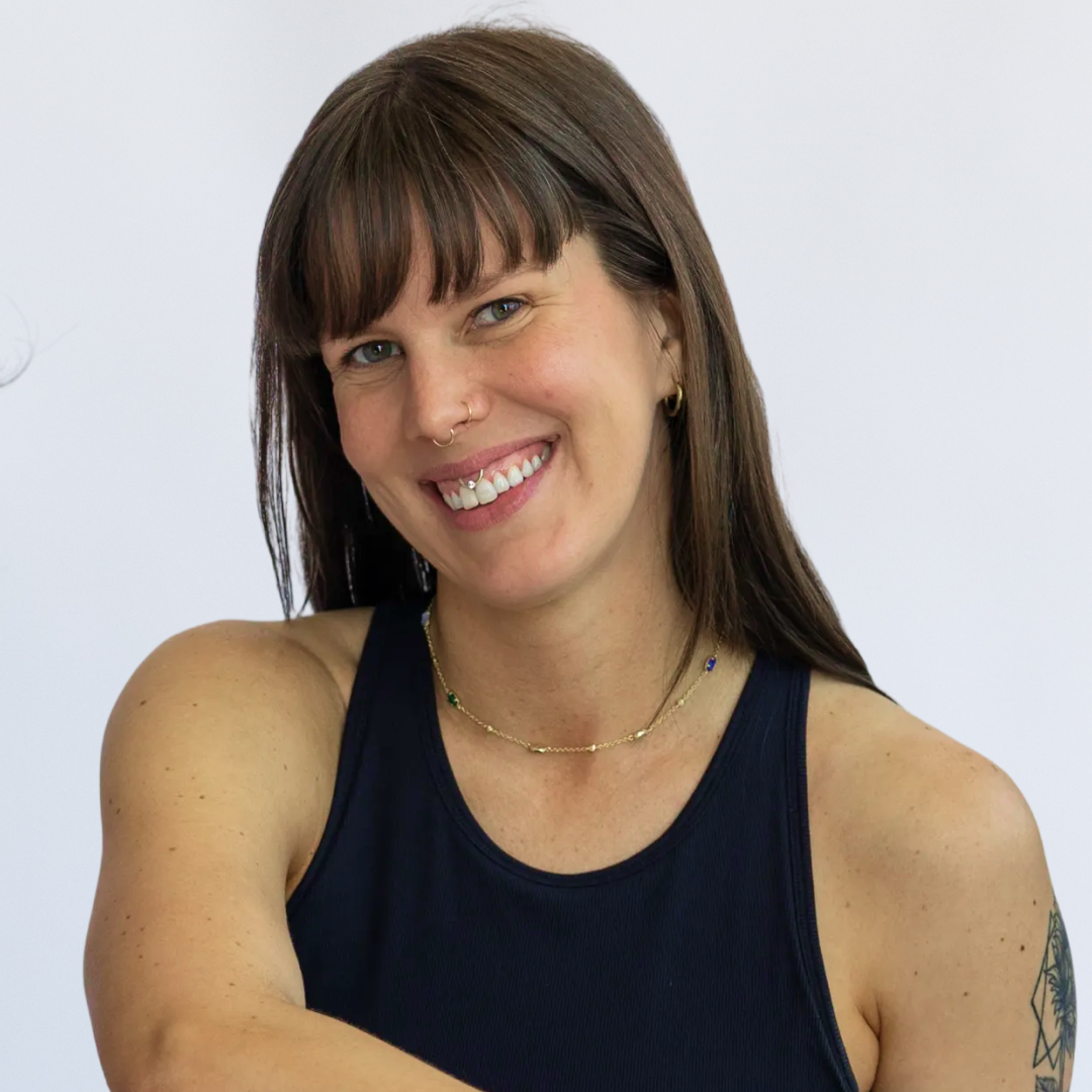 Woman with brown hair and tattoos smiling with arms crossed, wearing a black sleeveless top, against a plain white background.