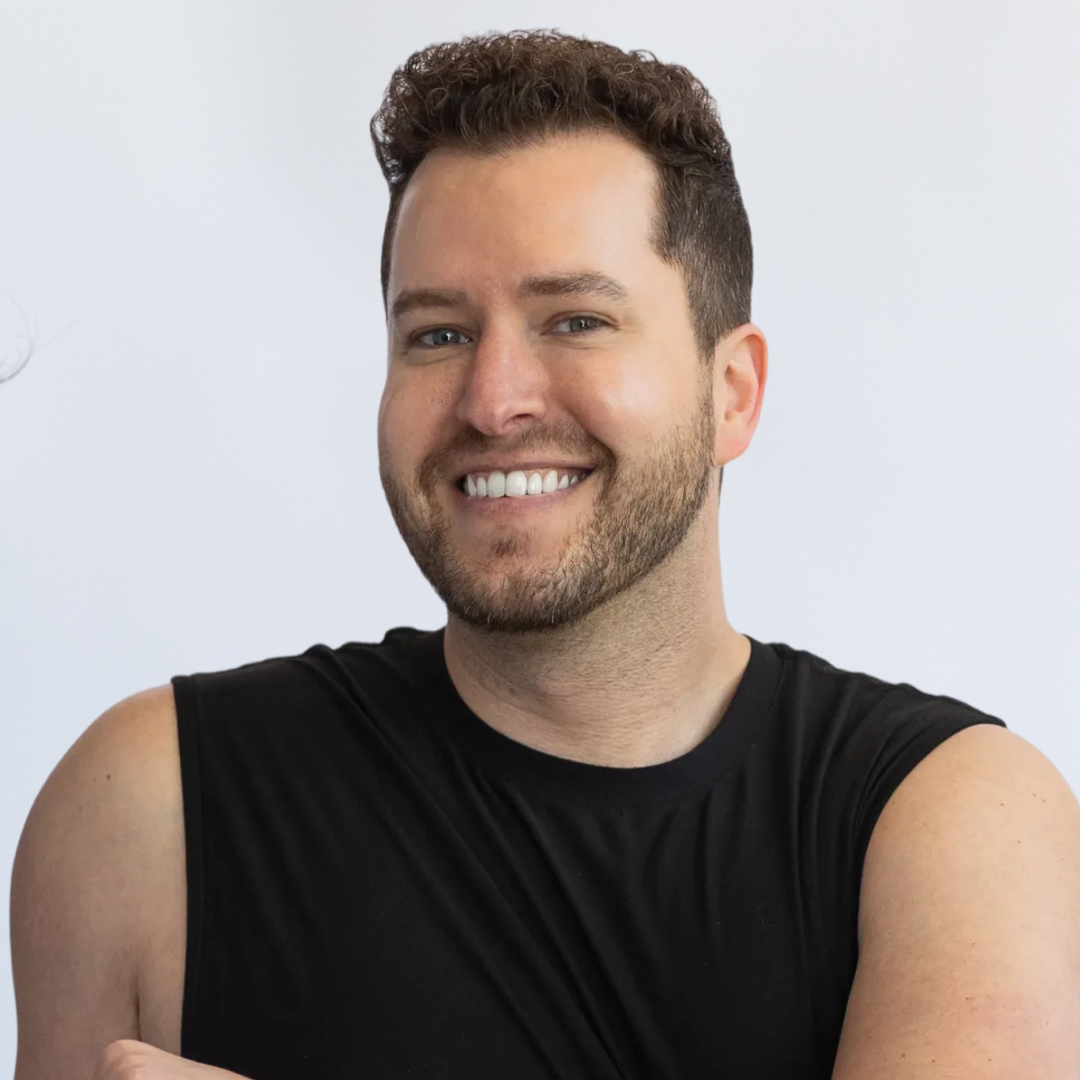 A smiling man with short curly hair and a beard, wearing a black sleeveless shirt, sitting against a plain white background.