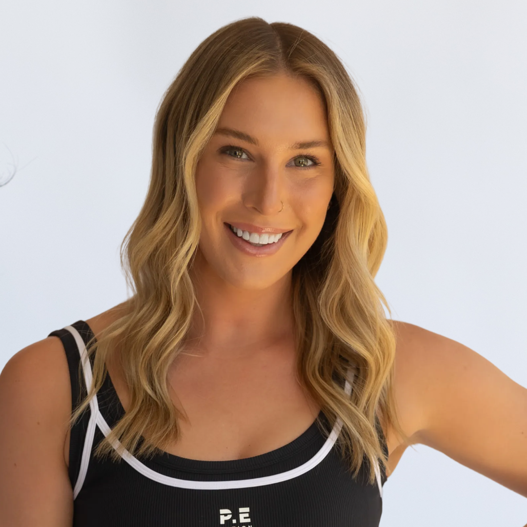 Portrait of a smiling woman with long blonde hair wearing a black sports bra and high-waisted black leggings, standing against a plain white background.