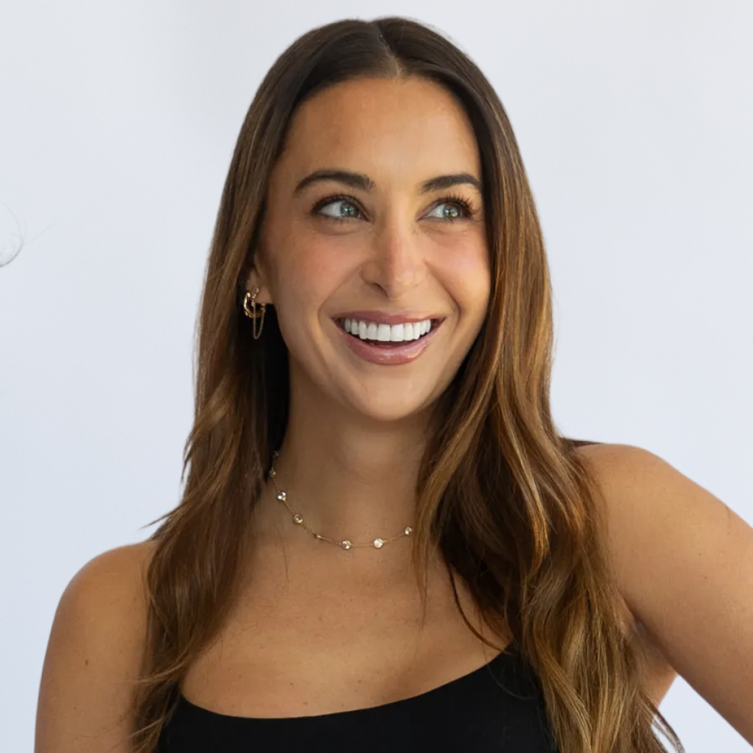 Woman with long brown hair in black sports bra and shorts holding dumbbells, standing indoors against a white background.
