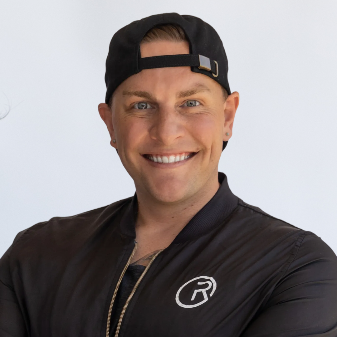 A smiling man with blue eyes, wearing a black jacket and a backward black cap, standing against a plain background.