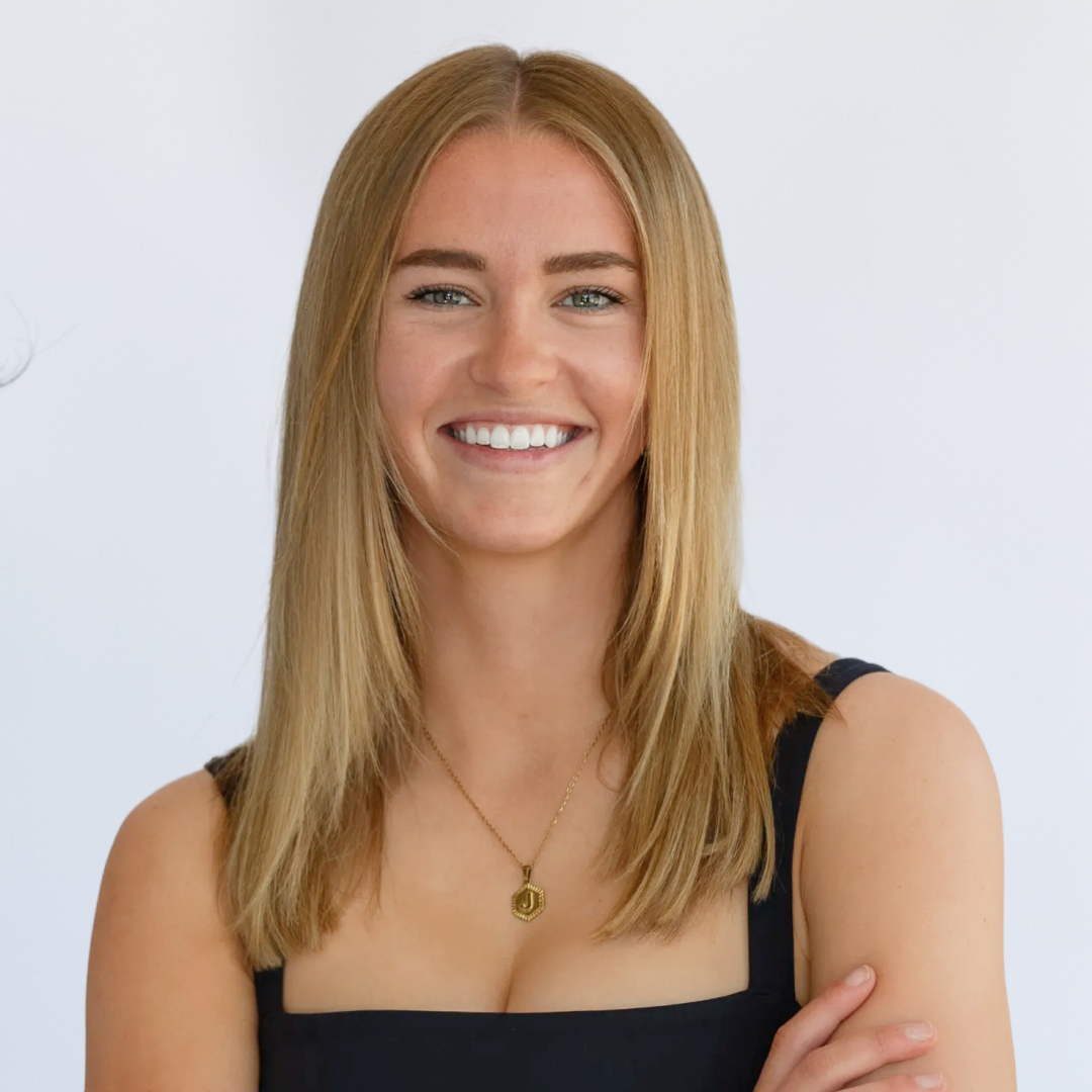 A smiling young woman with long blonde hair wearing a black sports bra and a gold necklace, sitting on a white background.
