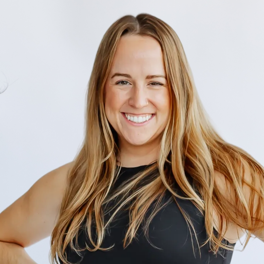 A woman smiling at the camera with long blonde hair, wearing a black sleeveless athletic top and gloves, standing indoors near a white wall.