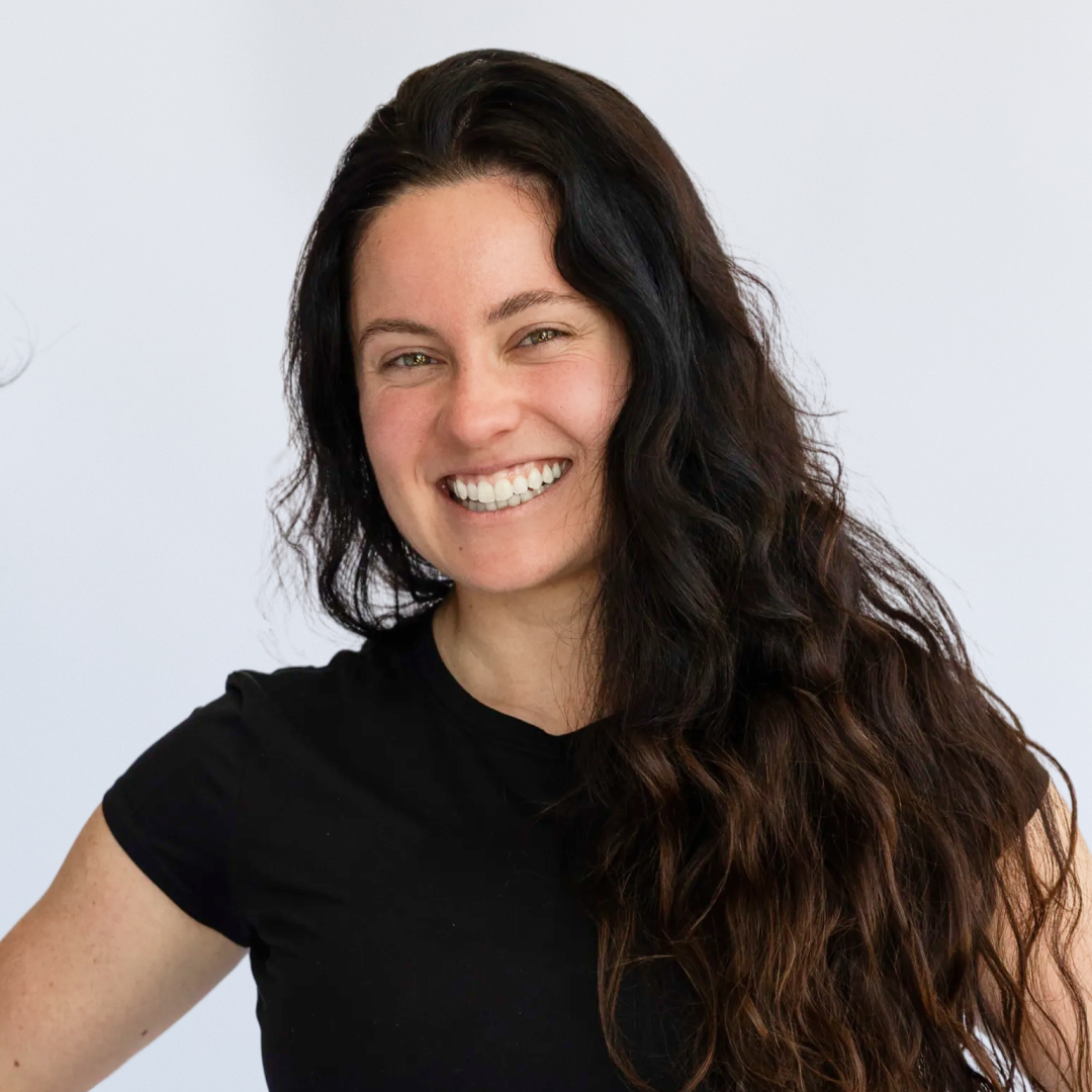 A young woman with long dark curly hair, smiling, wearing a black t-shirt and black pants, standing with hands on hips against a white background.